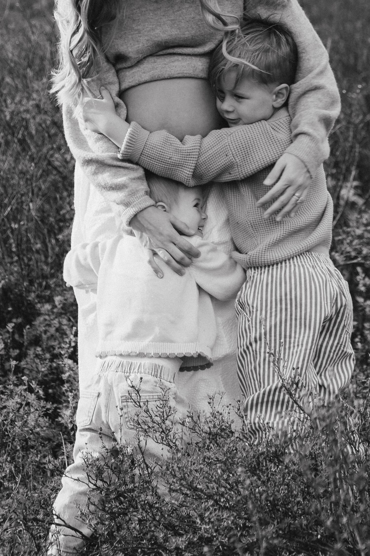 Black and white photo of pregnant woman being hugged by two young children in a field.