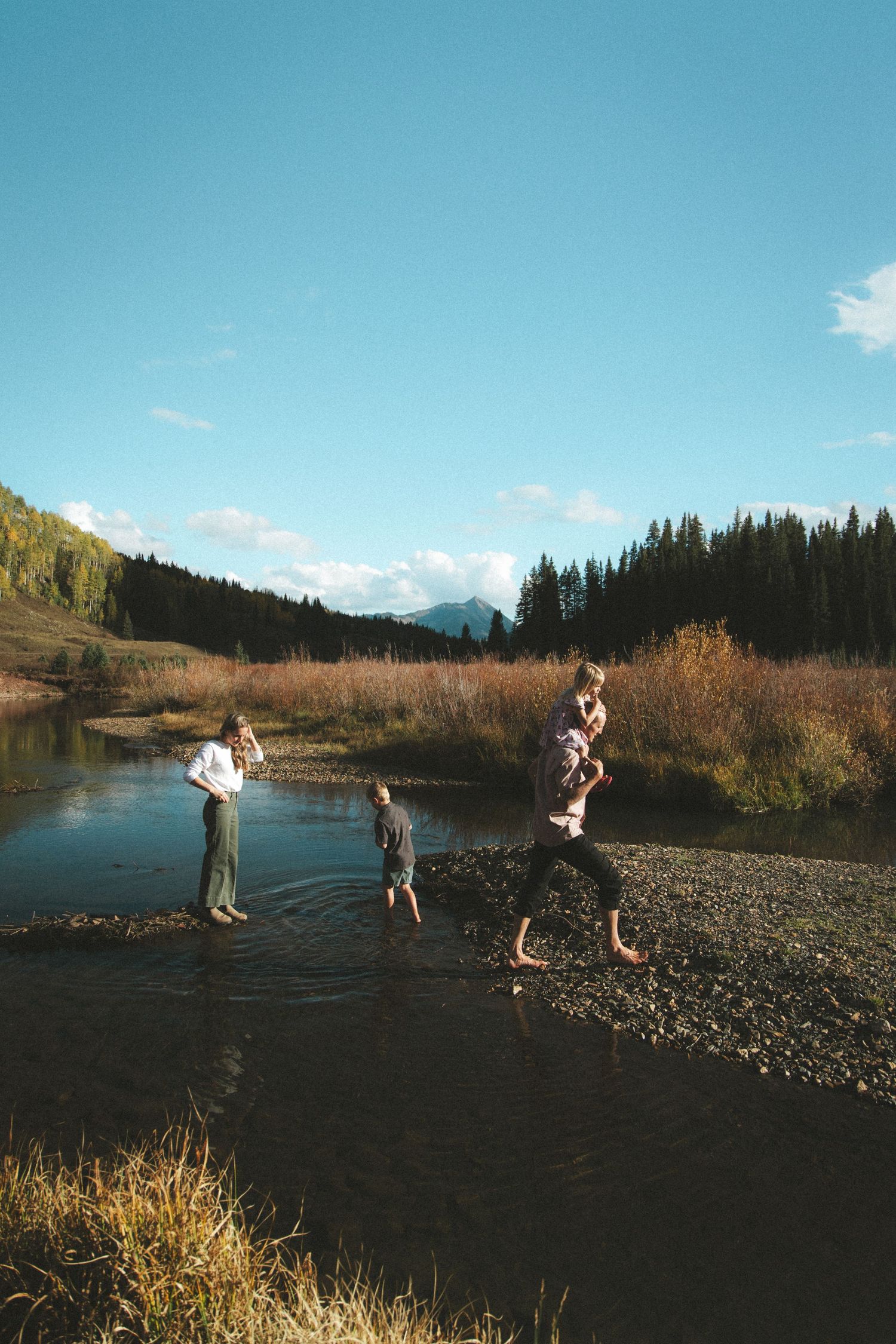 Family playing in a shallow mountain stream surrounded by autumn foliage and evergreen trees under a clear blue sky.