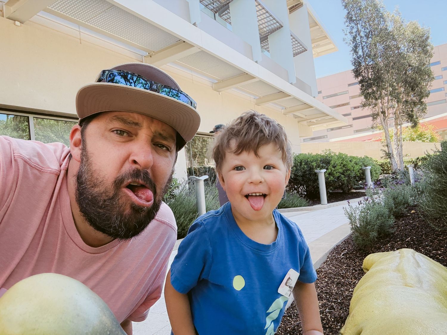 A man in a snapback hat and a young boy in a blue shirt smile at the camera outdoors near pumpkins.