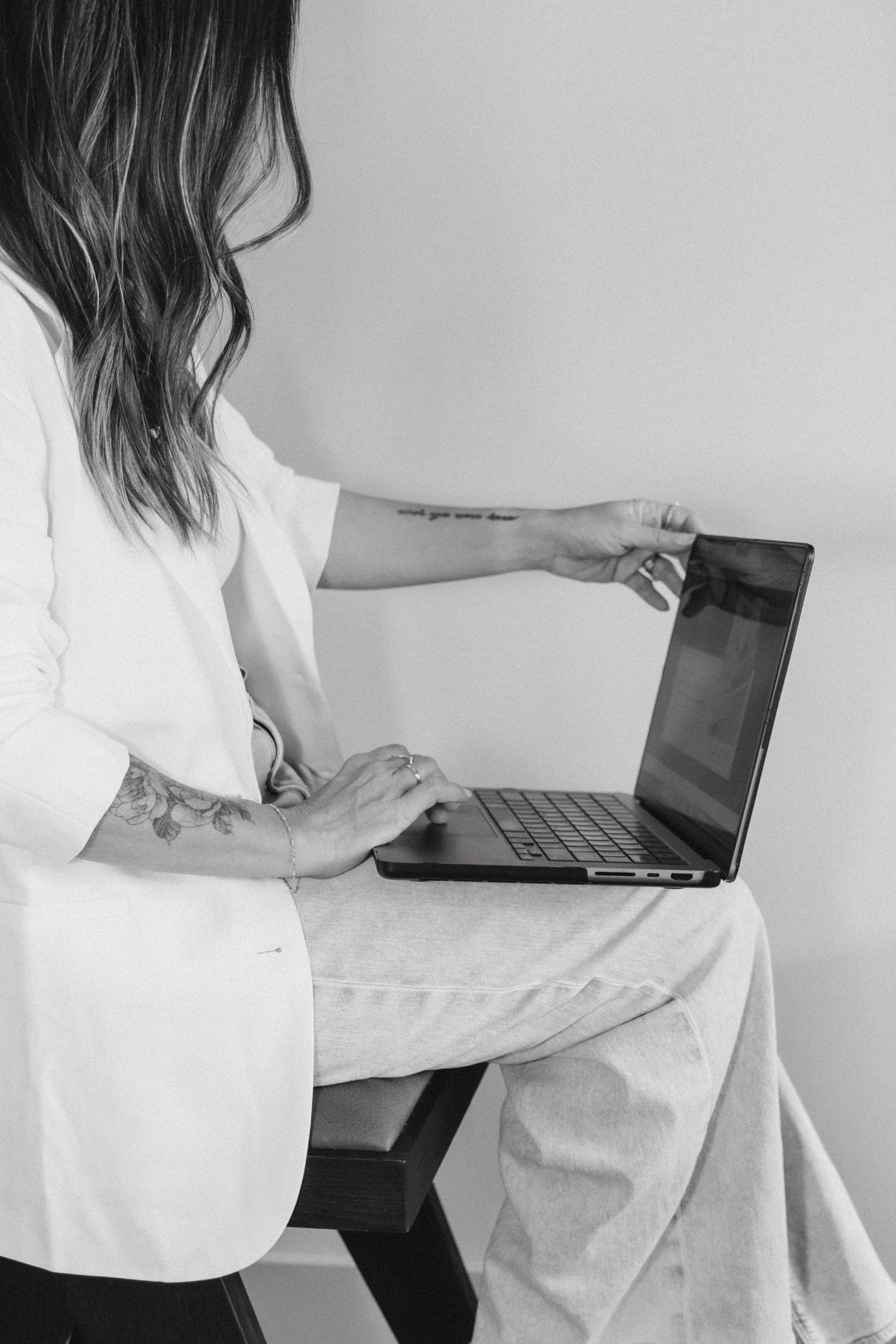 Black and white photo of a tattooed woman in white blazer sitting and working on a laptop computer.