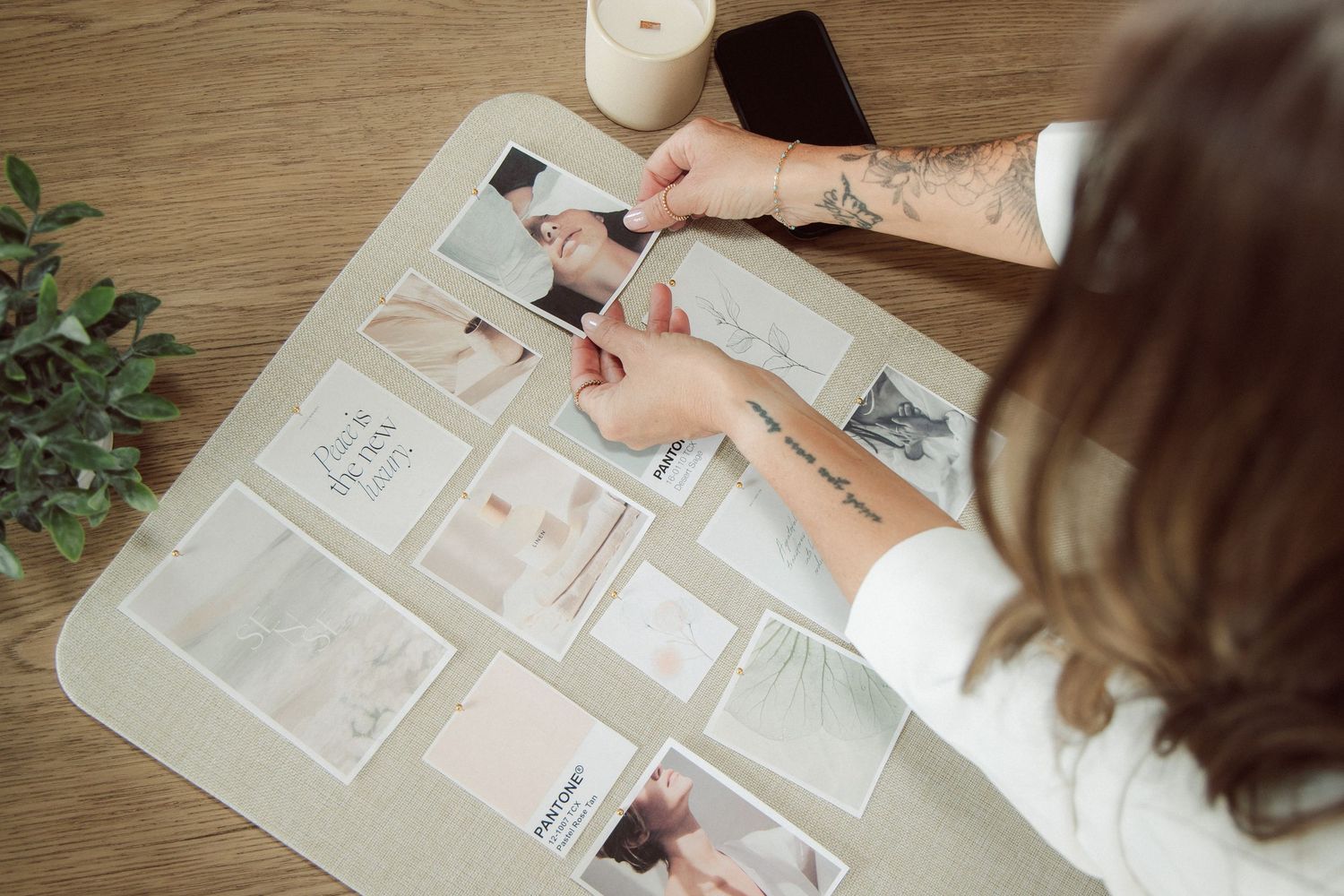 A tattooed woman arranges photos and cards on a desk while creating a mood board near a candle and smartphone.