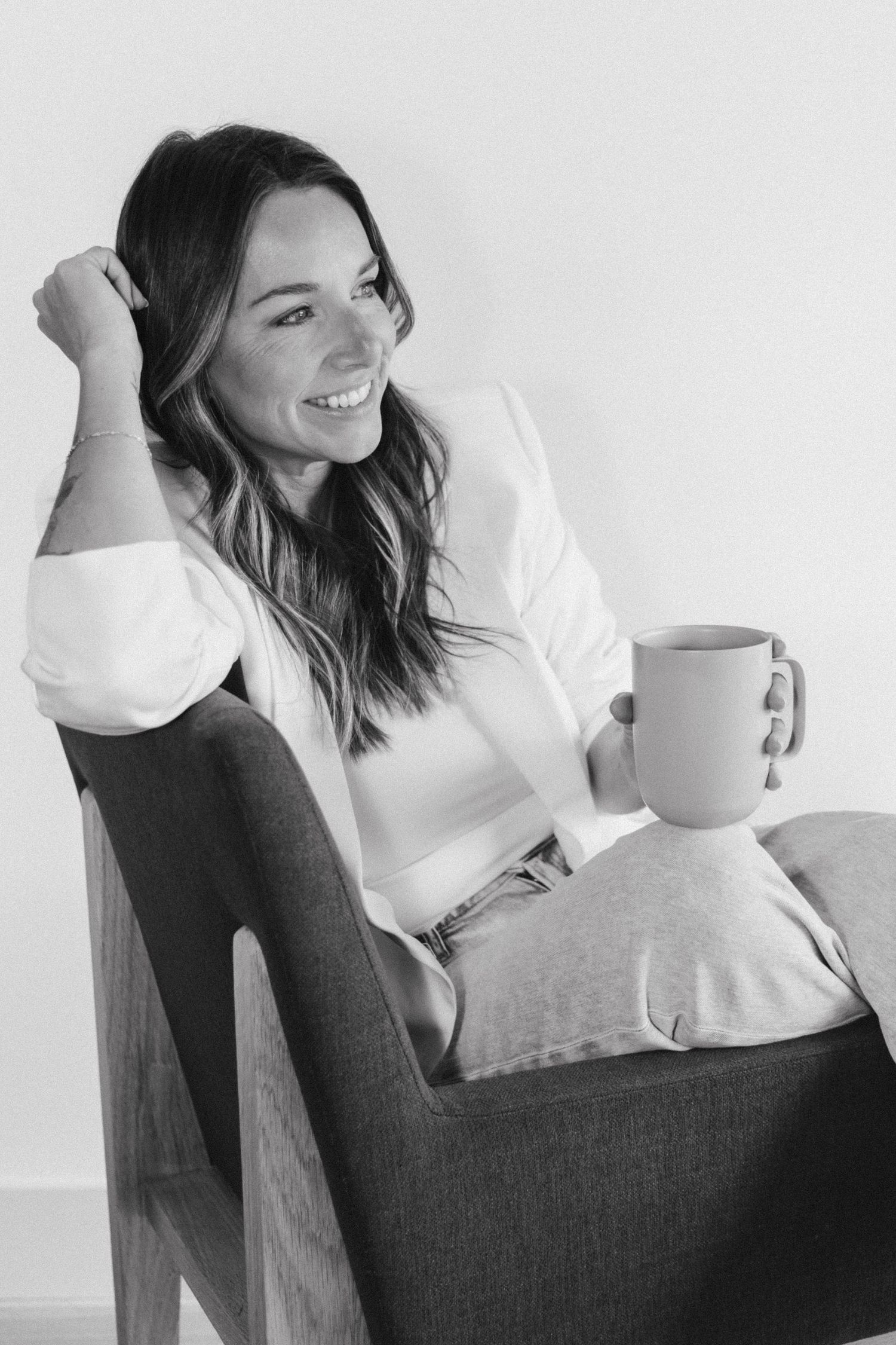 Smiling woman sitting in armchair holding coffee mug, wearing white shirt and jeans, black and white portrait photo.
