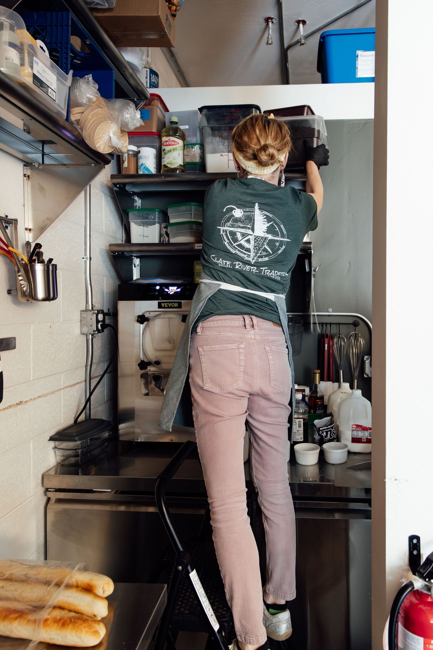 Woman in green t-shirt standing on step stool reaching upper shelves in a busy restaurant kitchen.
