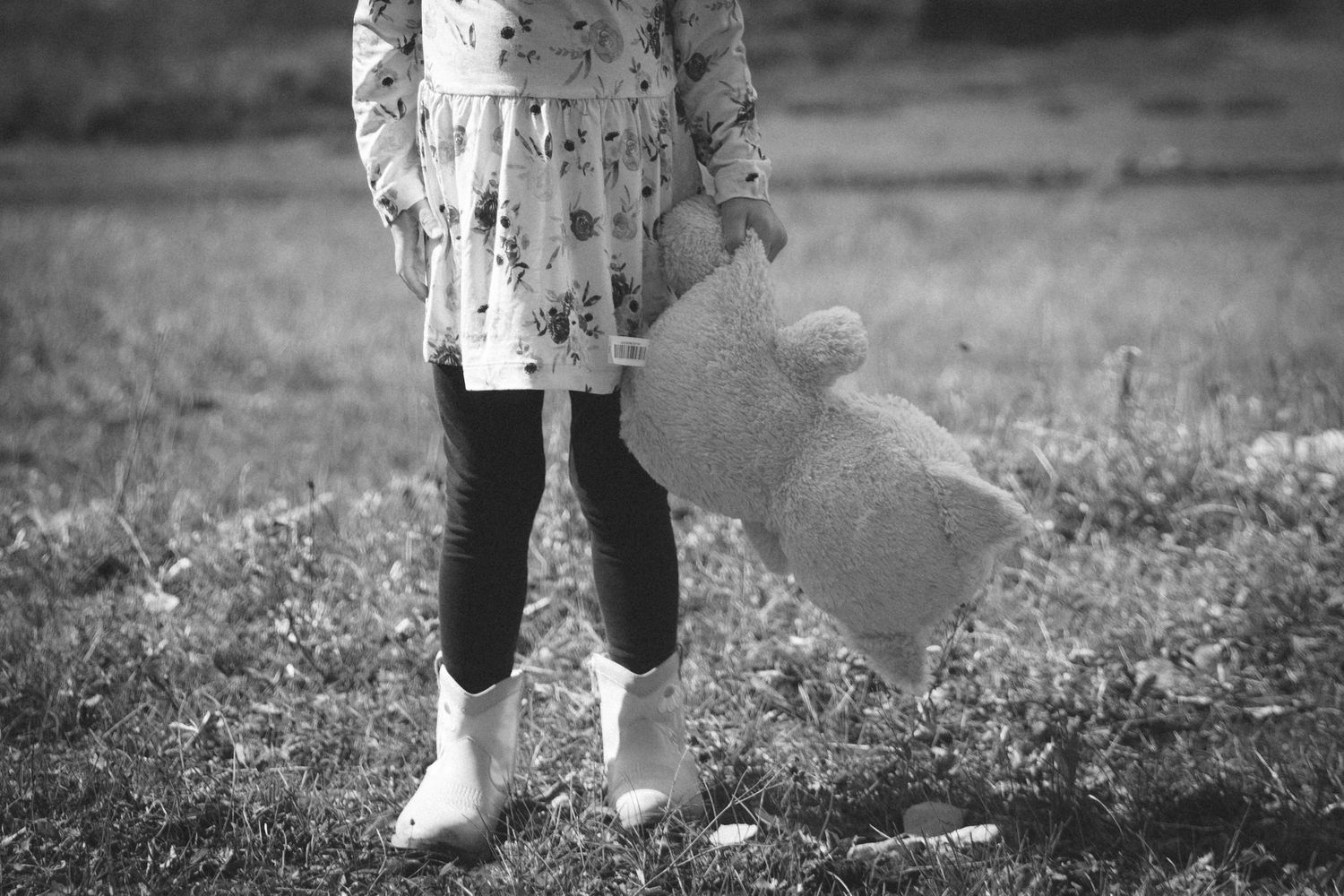 Black and white photo of a young girl holding a teddy bear while standing in a grassy field wearing boots.