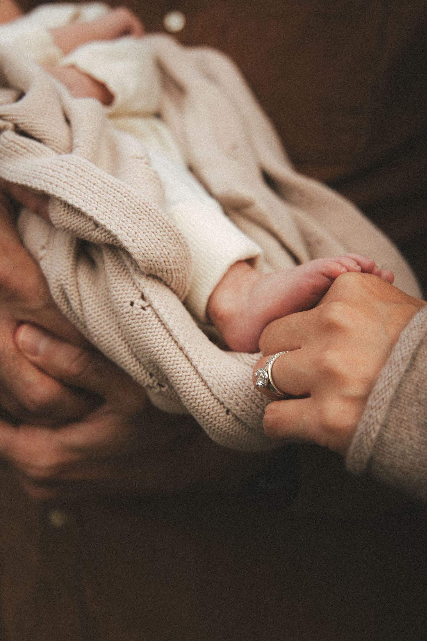 Adult hands in warm brown clothing gently hold a newborn wrapped in a cream knit blanket, with a small bare foot and a silver ring visible.