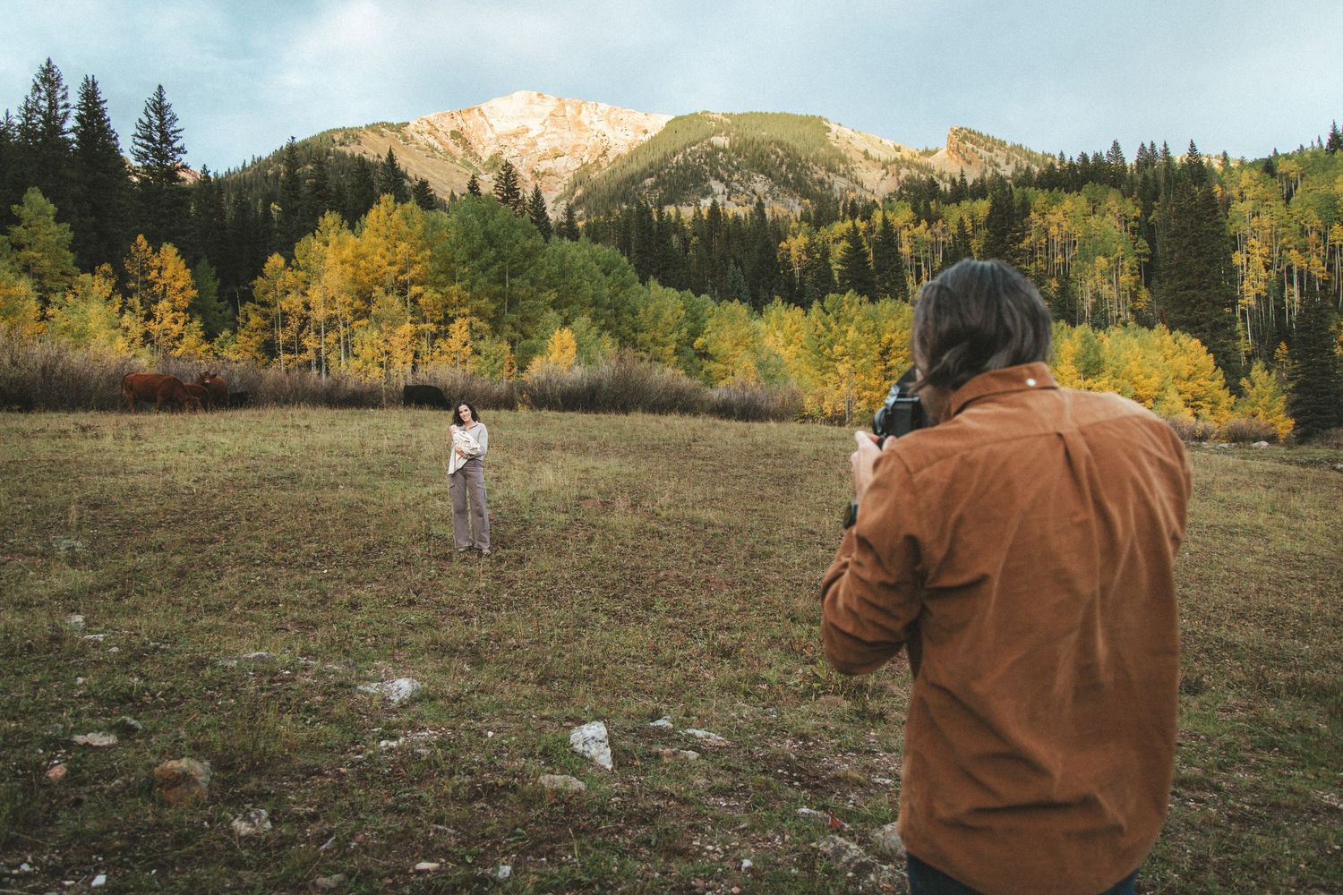 Photographer in a rust-colored jacket shoots a person standing in a mountain meadow surrounded by golden autumn aspens and evergreen trees.