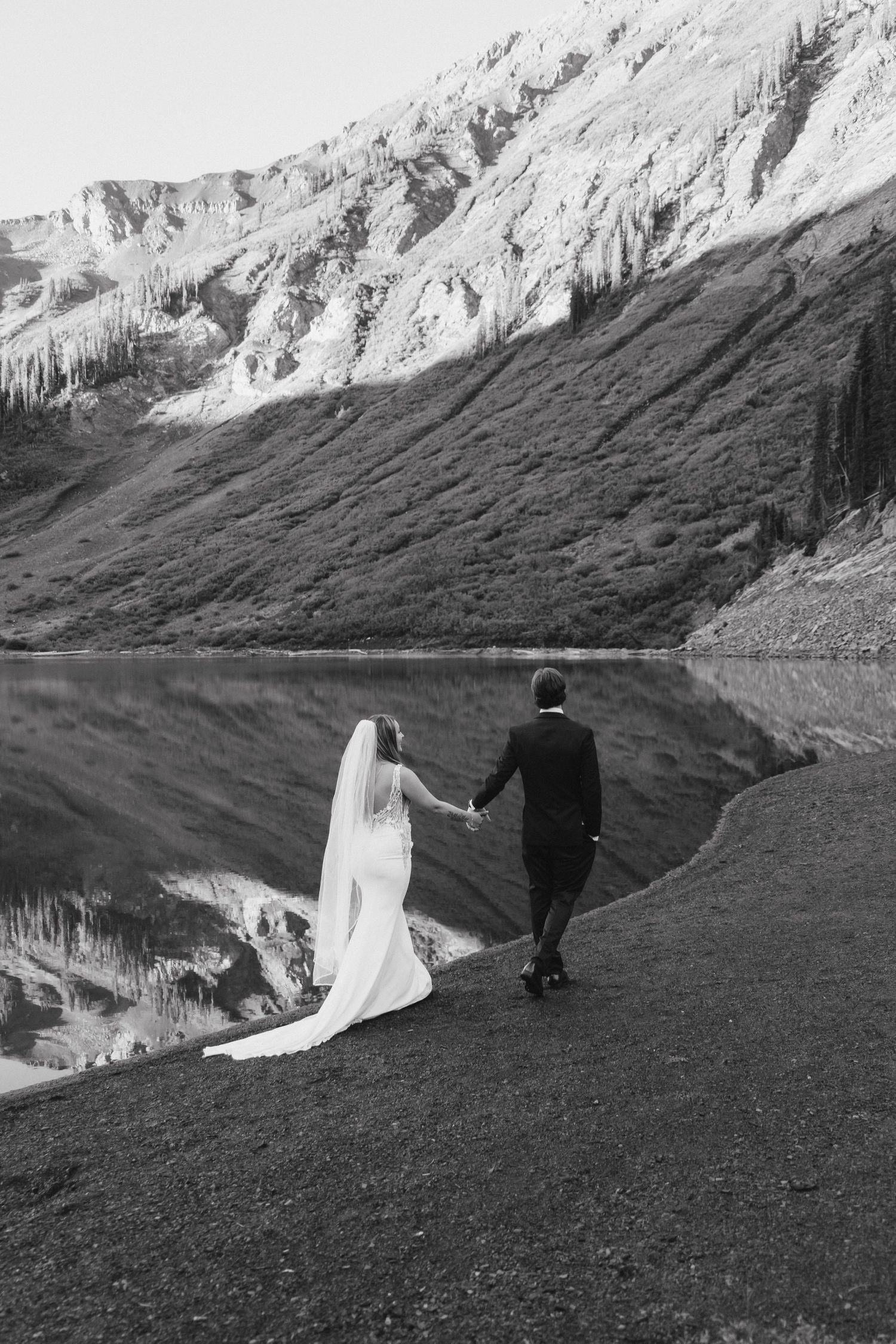 Black and white photo of a bride and groom holding hands walking along a mountain lake shoreline.