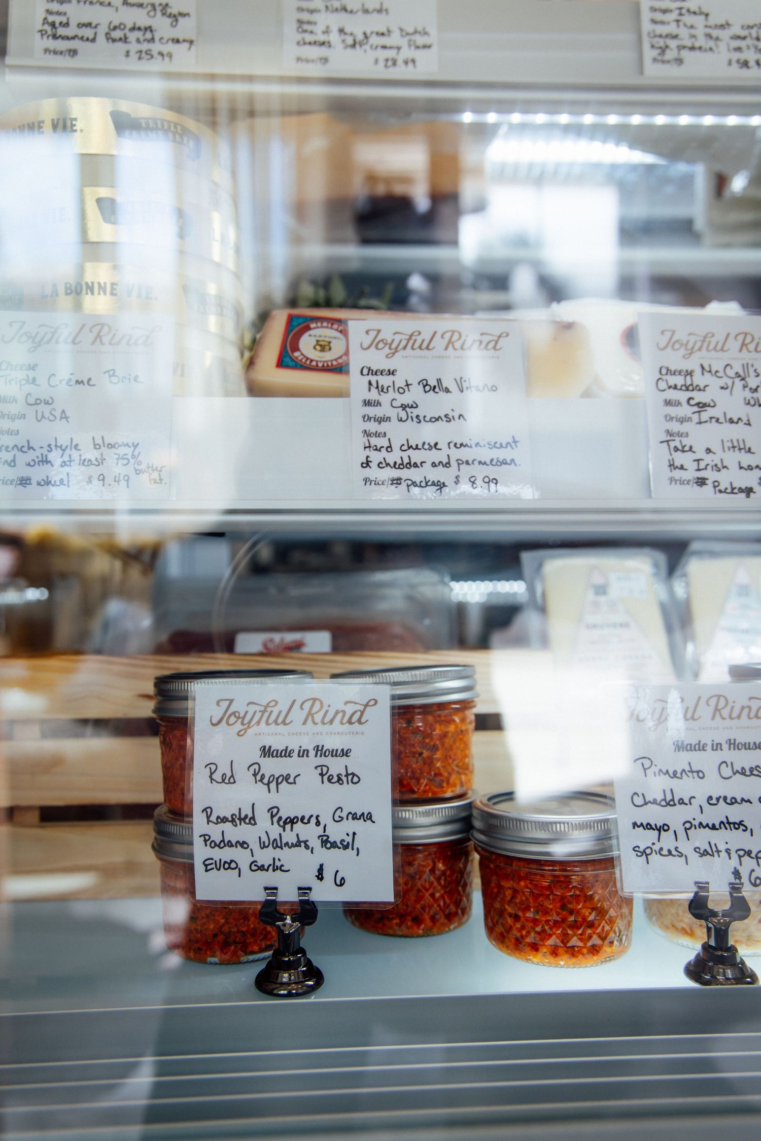 Glass display case at Joyful Rind cheese shop featuring red pepper pesto jars and artisan cheese selections with handwritten labels.