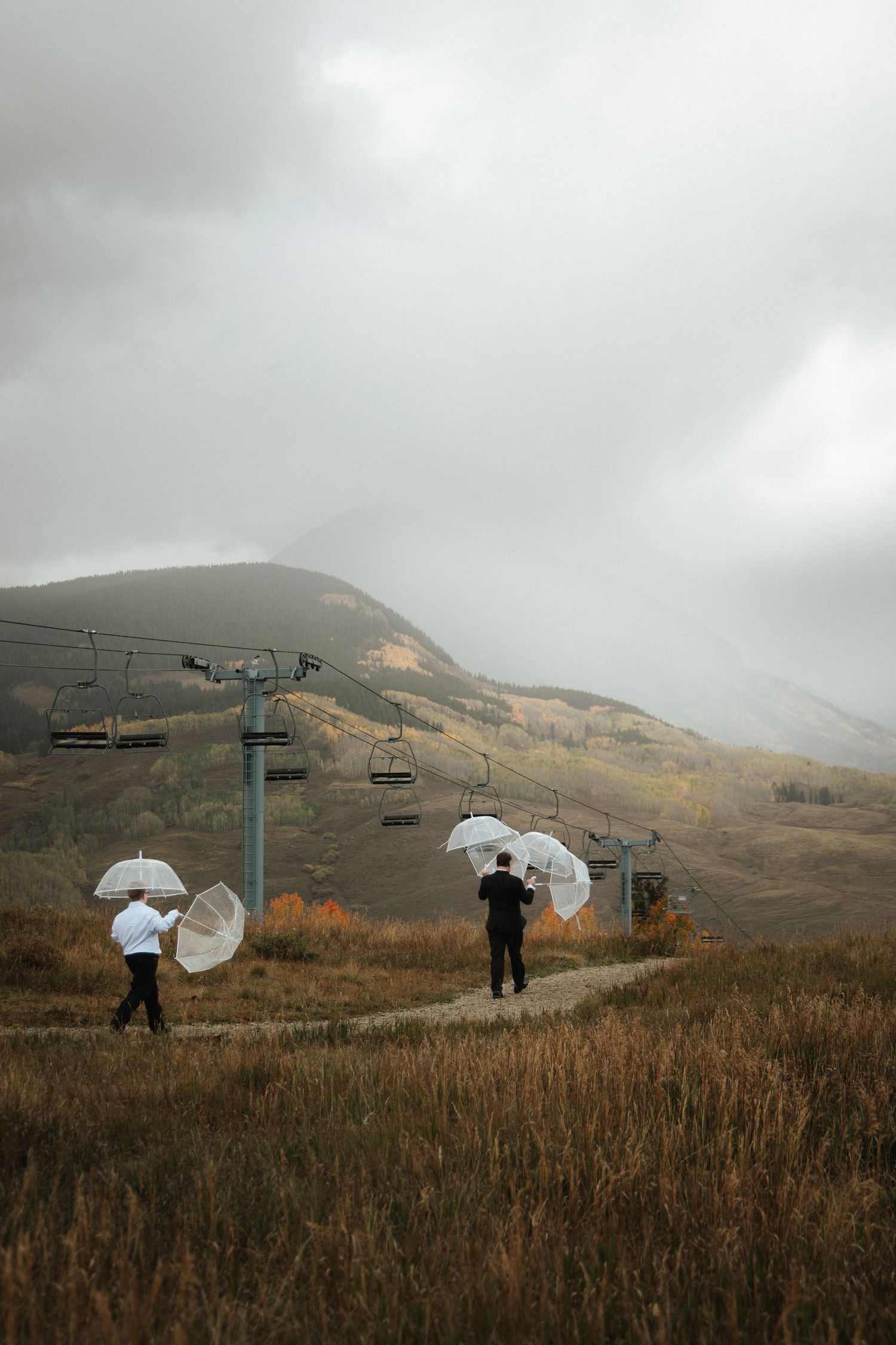 Two people in formal attire walk along a dirt path carrying clear umbrellas near a ski lift on a moody, overcast autumn mountain hillside.