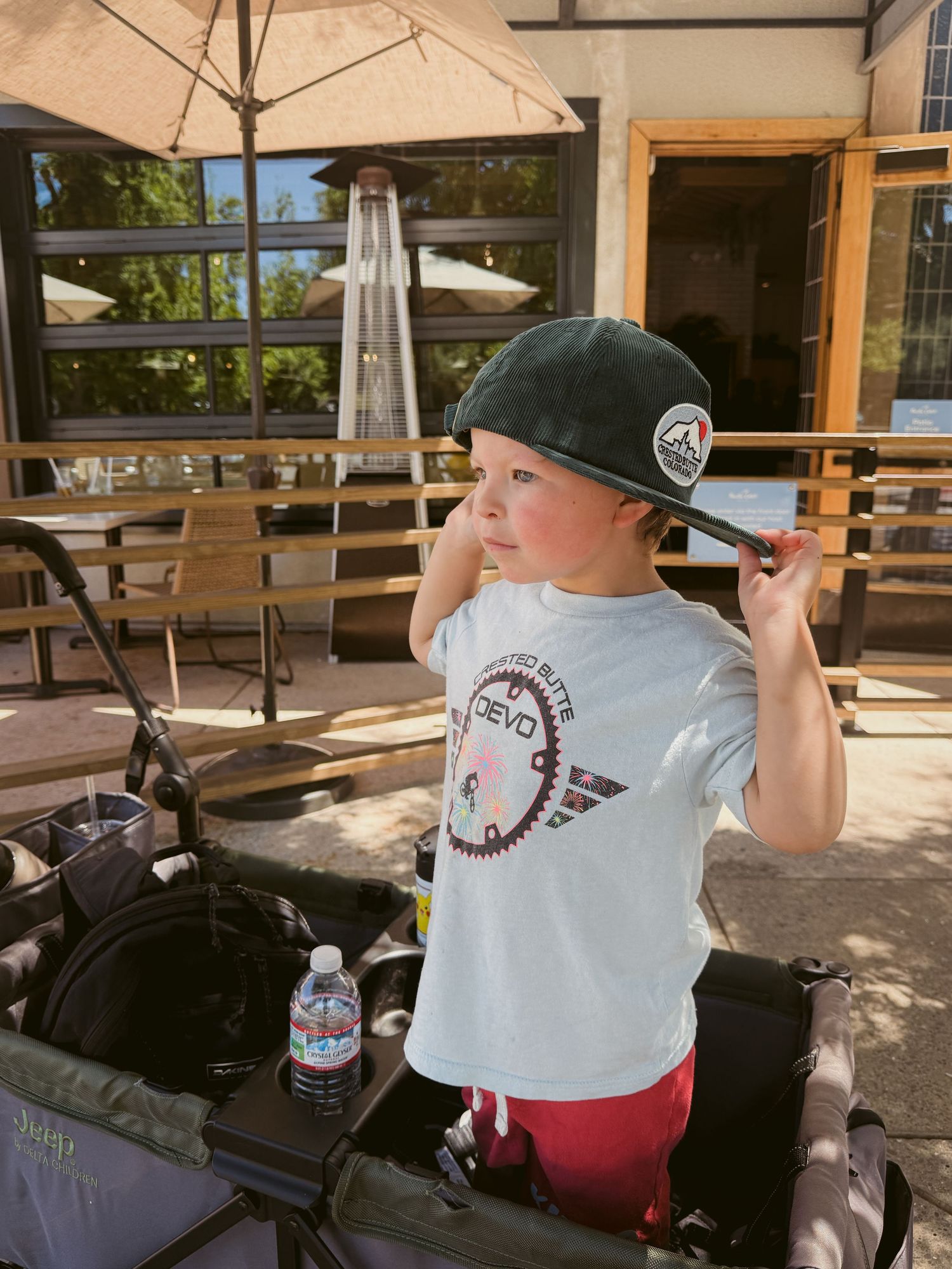 Young child wearing a dark snapback cap and white graphic tee stands outdoors near a stroller on a sunny day.
