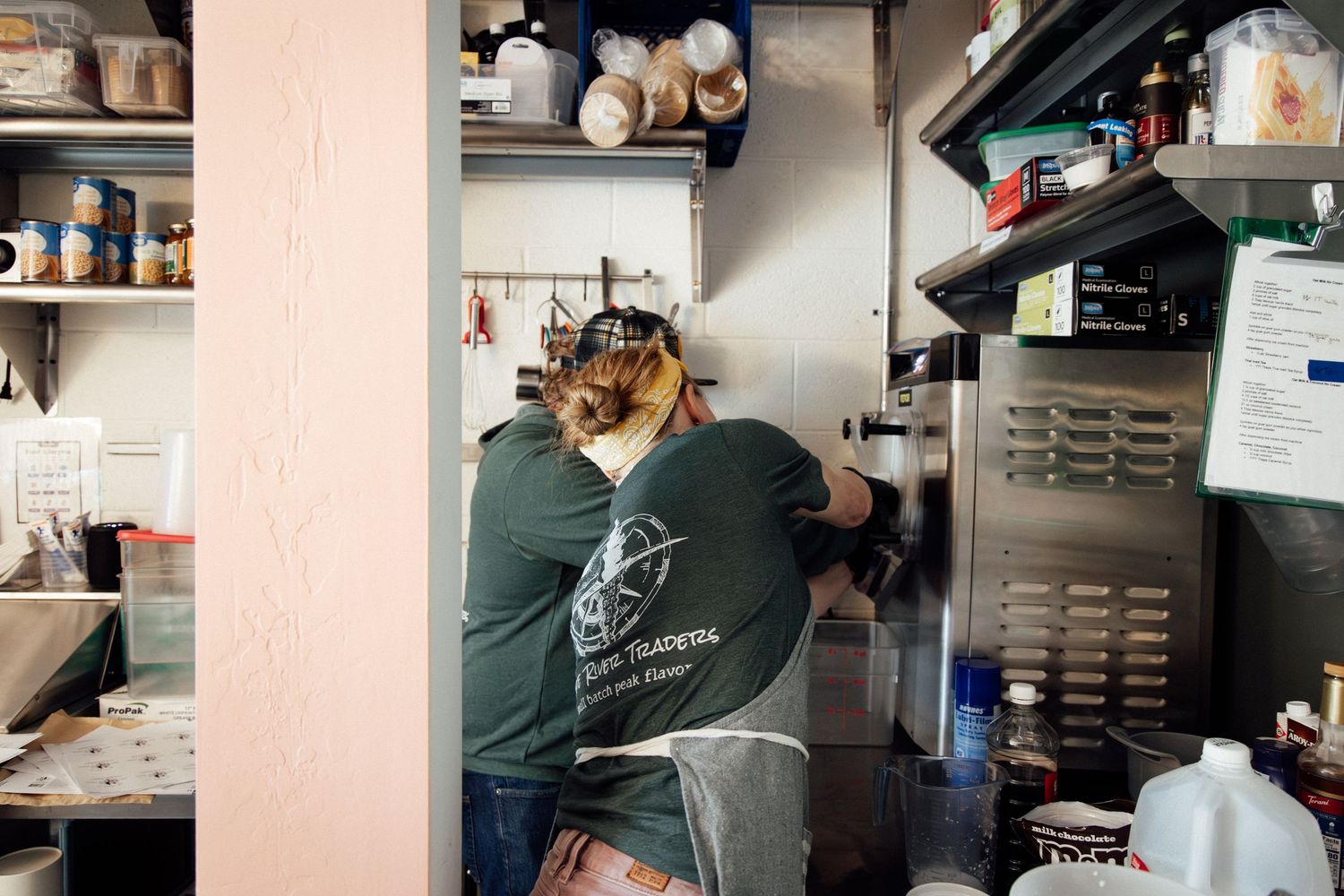 A kitchen worker in a green shirt prepares food at a commercial kitchen station with shelves and equipment.