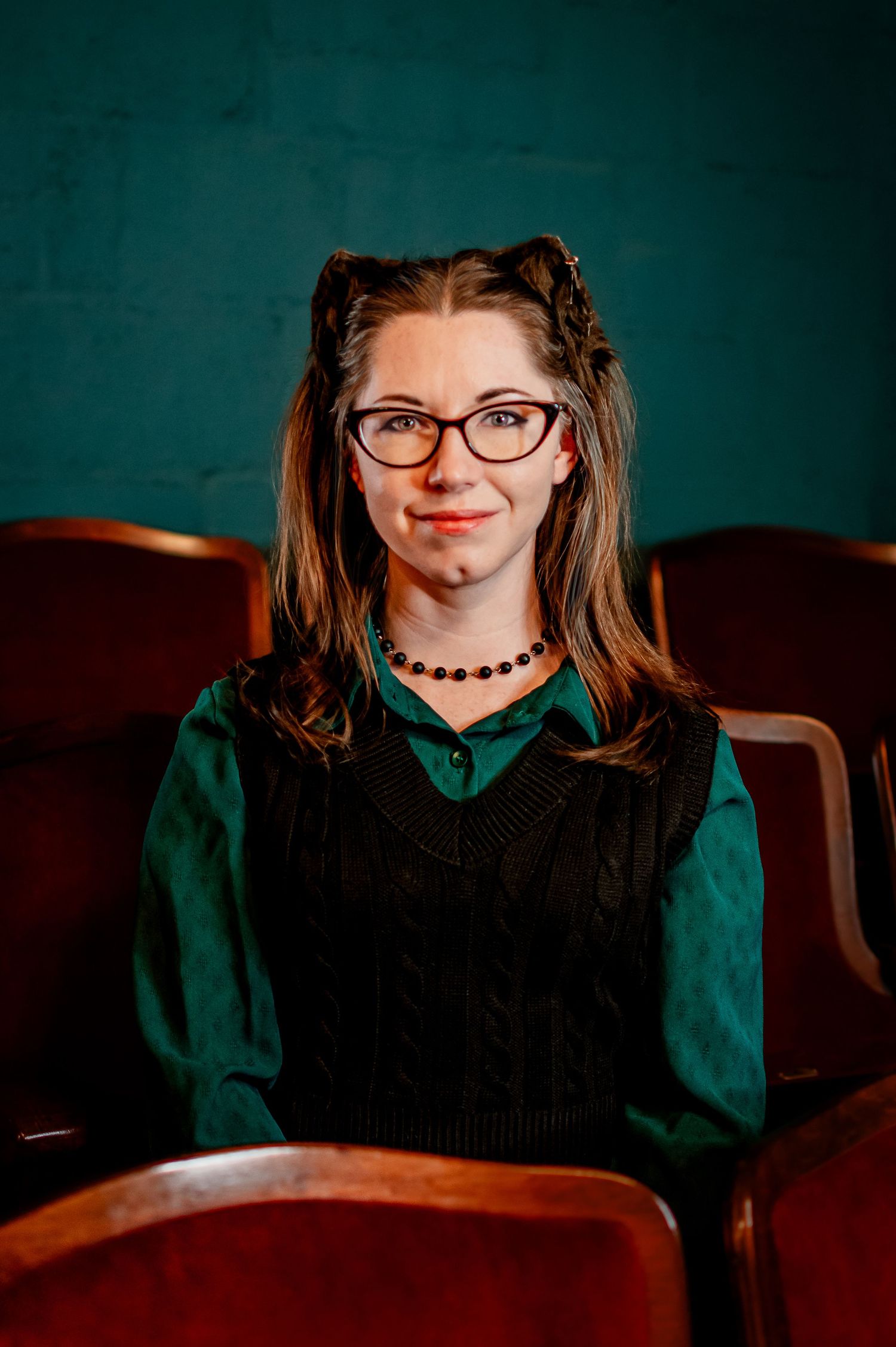 Retro-style portrait in a theater shows woman with cat ears wearing glasses, green shirt and black vest against teal background.