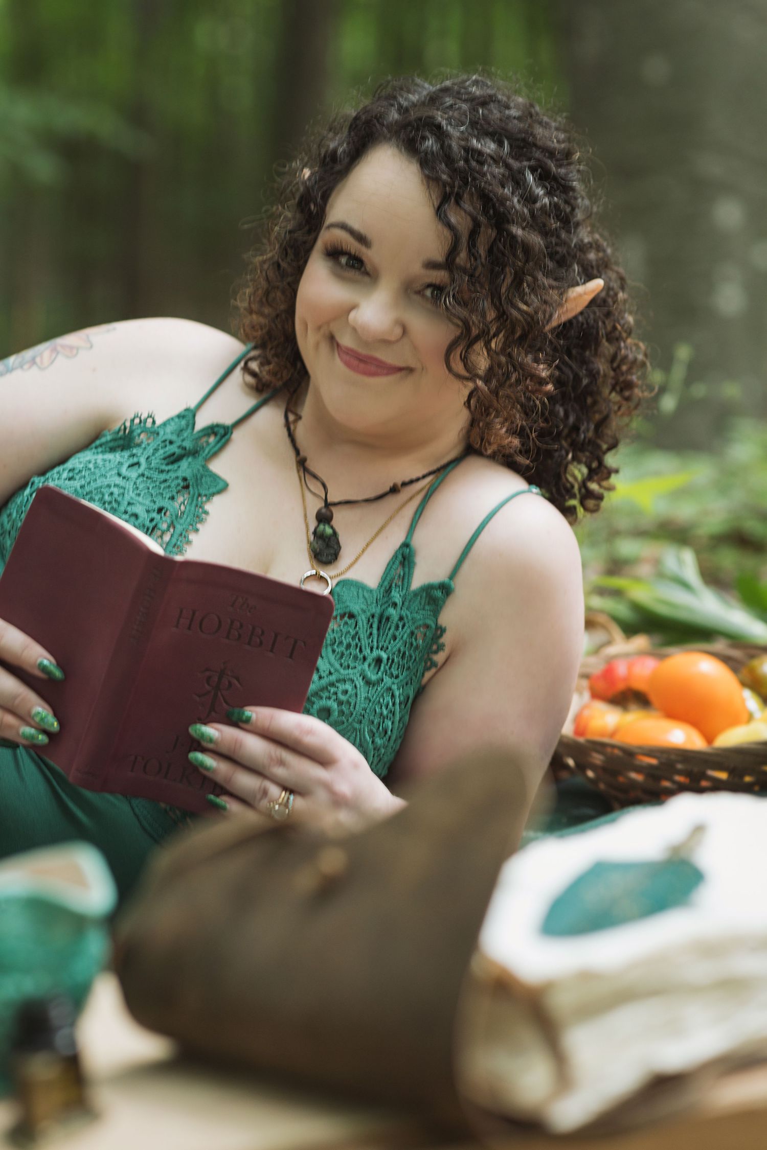 Fantasy woodland cosplay scene with reader in emerald lace holding leather bound The Hobbit book among forest foliage.