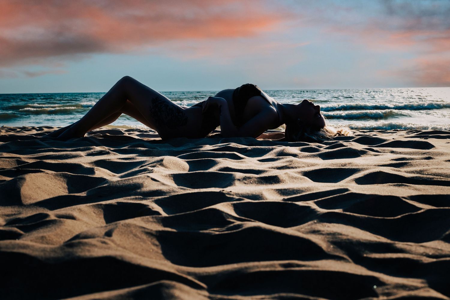 A silhouette lies across rippling beach sand at sunset with choppy ocean waves and pink clouds in the background.