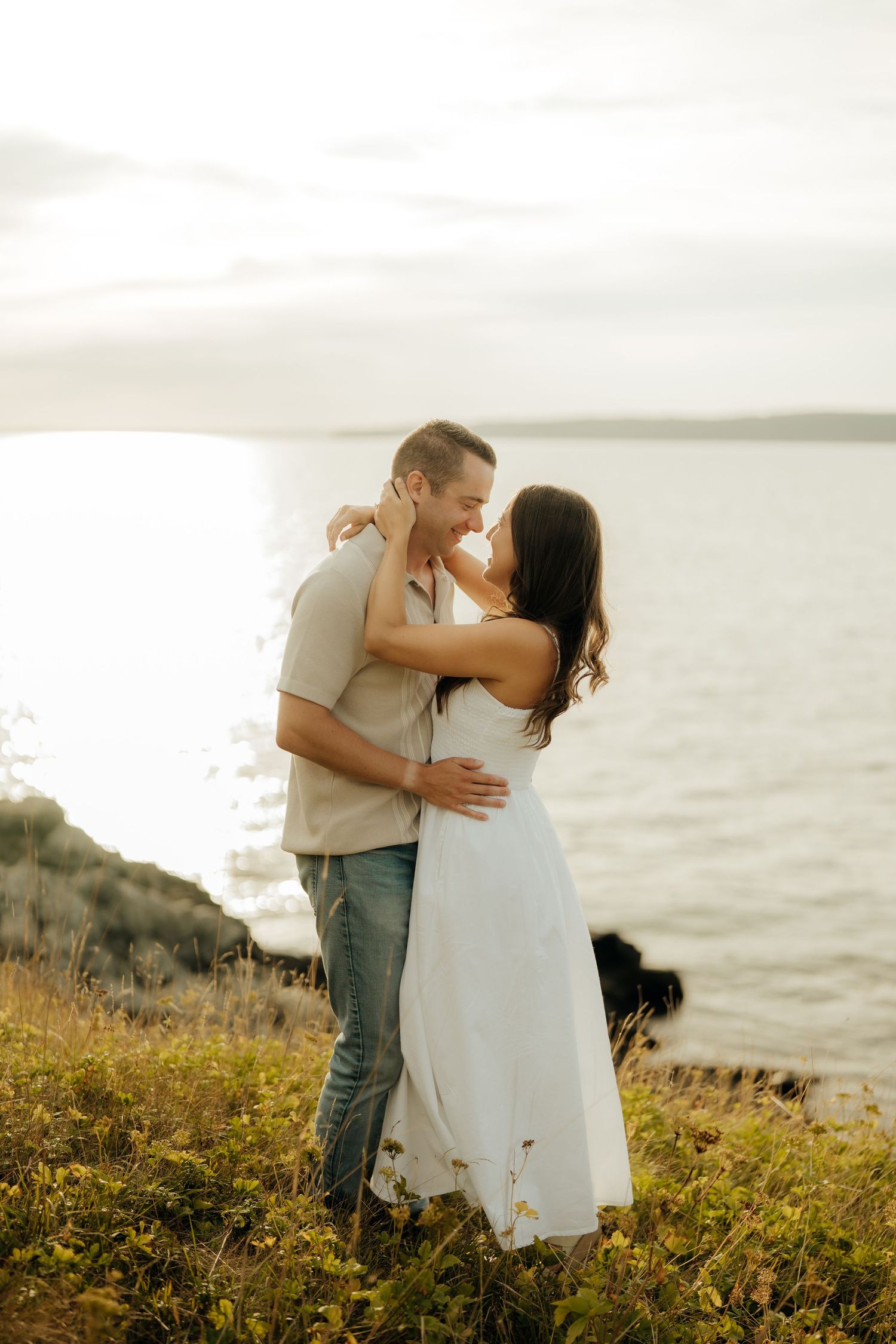 Romantic couple embraces on a coastal cliff with yellow wildflowers and ocean view at sunset.