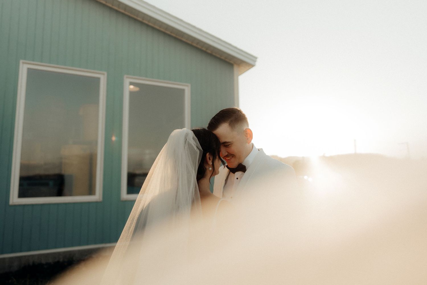A bride in a white wedding dress and veil is photographed in soft window light against a light-colored wall.