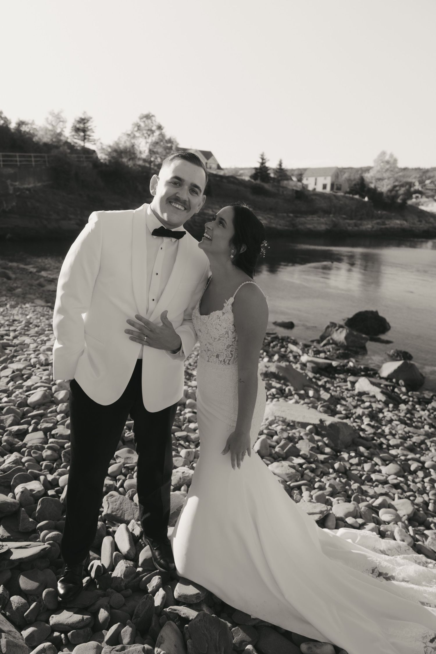 A couple in wedding attire poses together on rocky terrain near a waterfront during their romantic outdoor photoshoot.