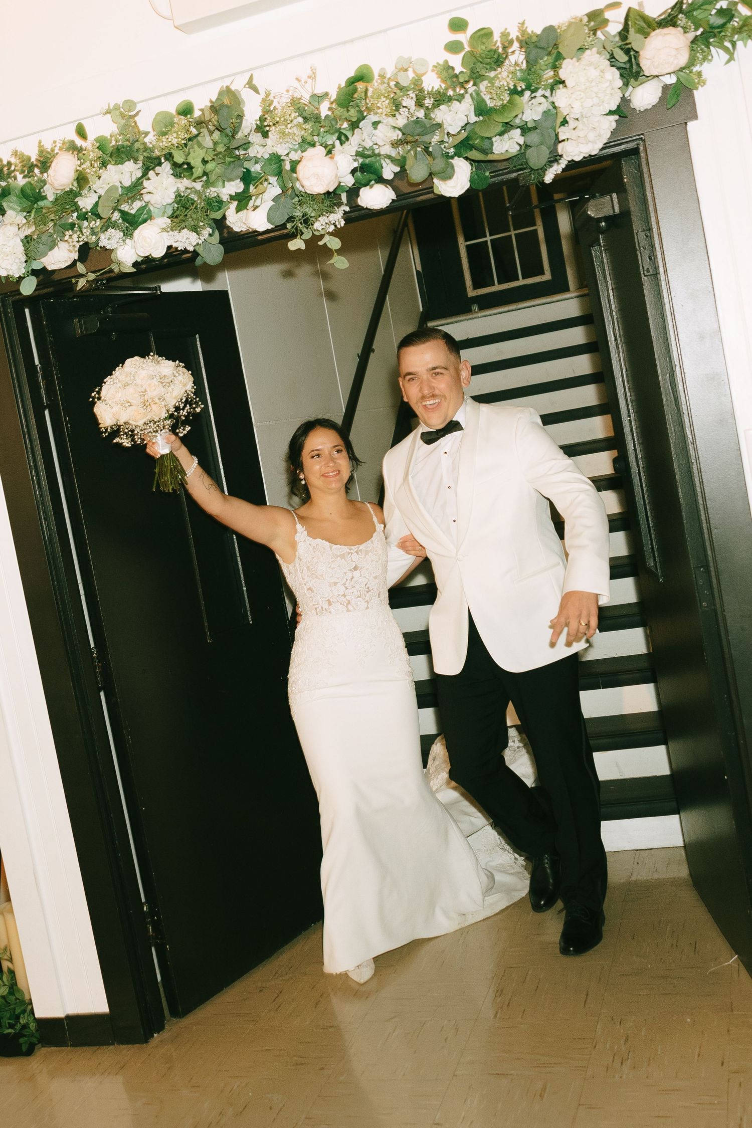 A couple in wedding attire poses by a staircase under an elegant floral arch with white blooms and greenery.
