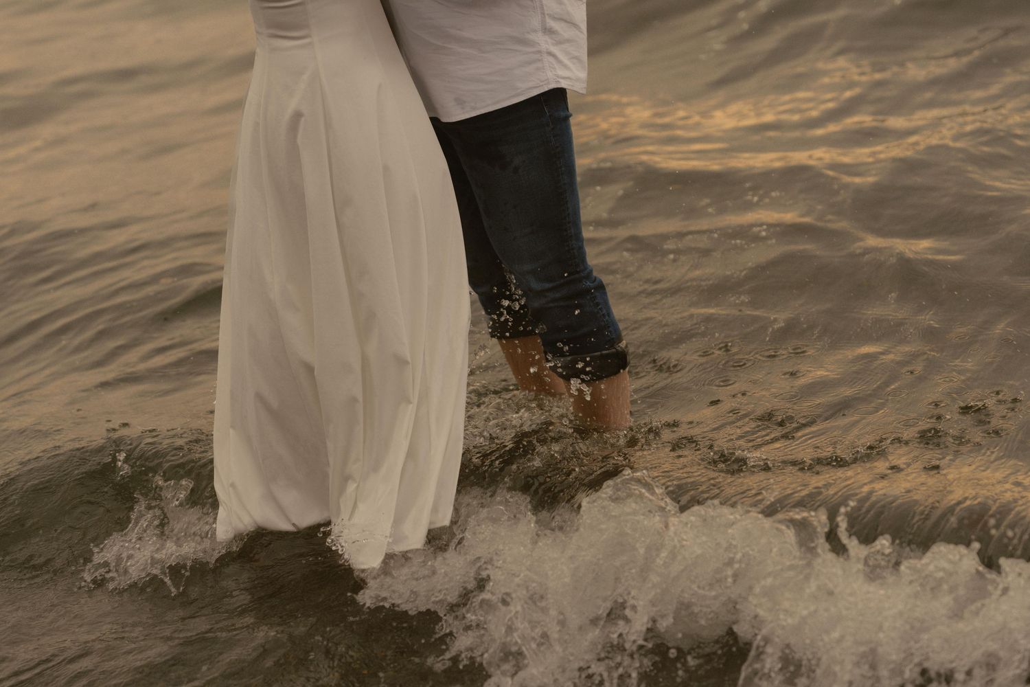 Two pairs of legs in white and dark clothing stand in shallow water as waves splash around their feet.