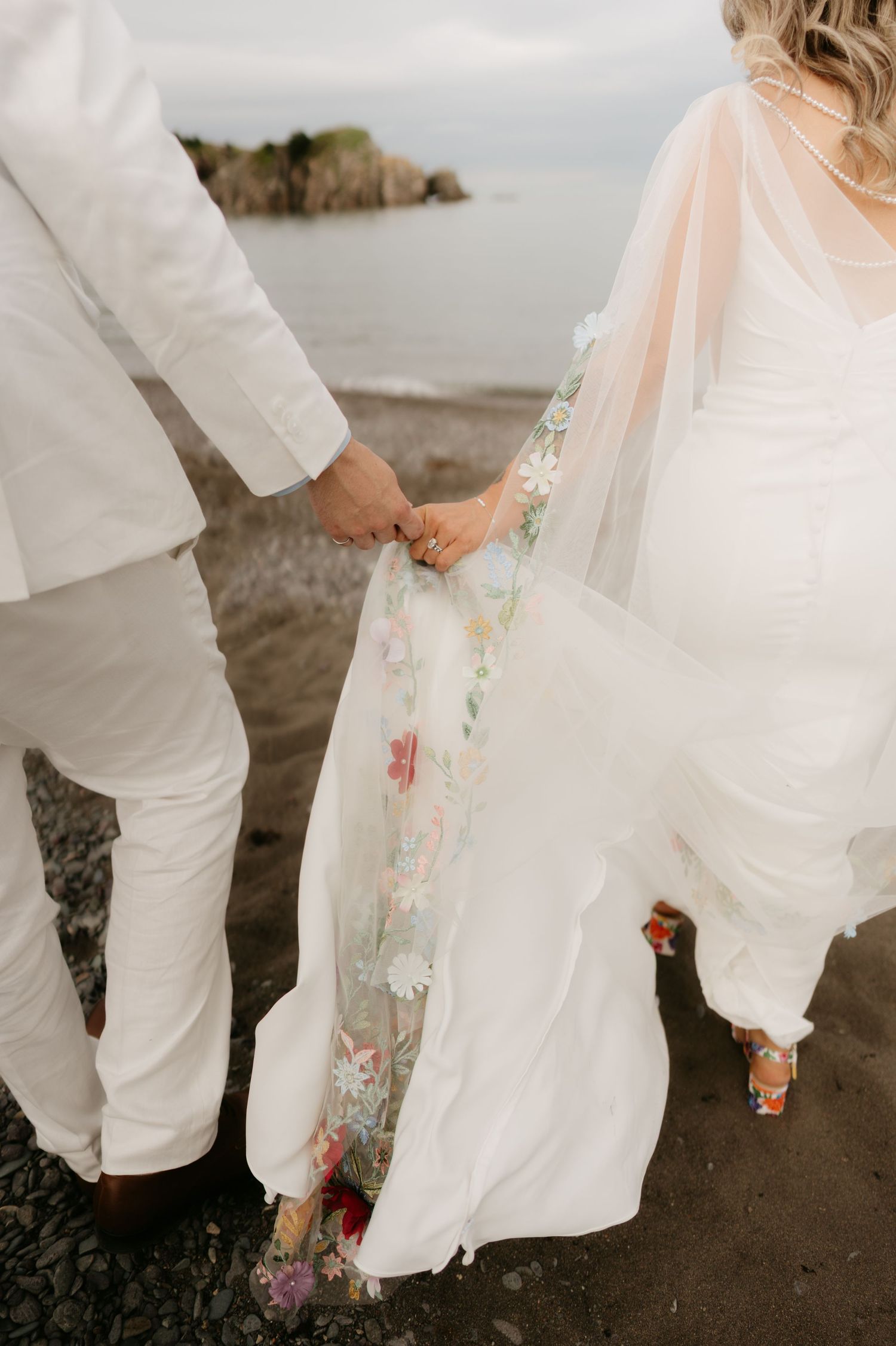 Bride and groom holding hands on rocky beach, bride wearing floral embroidered veil and white gown.