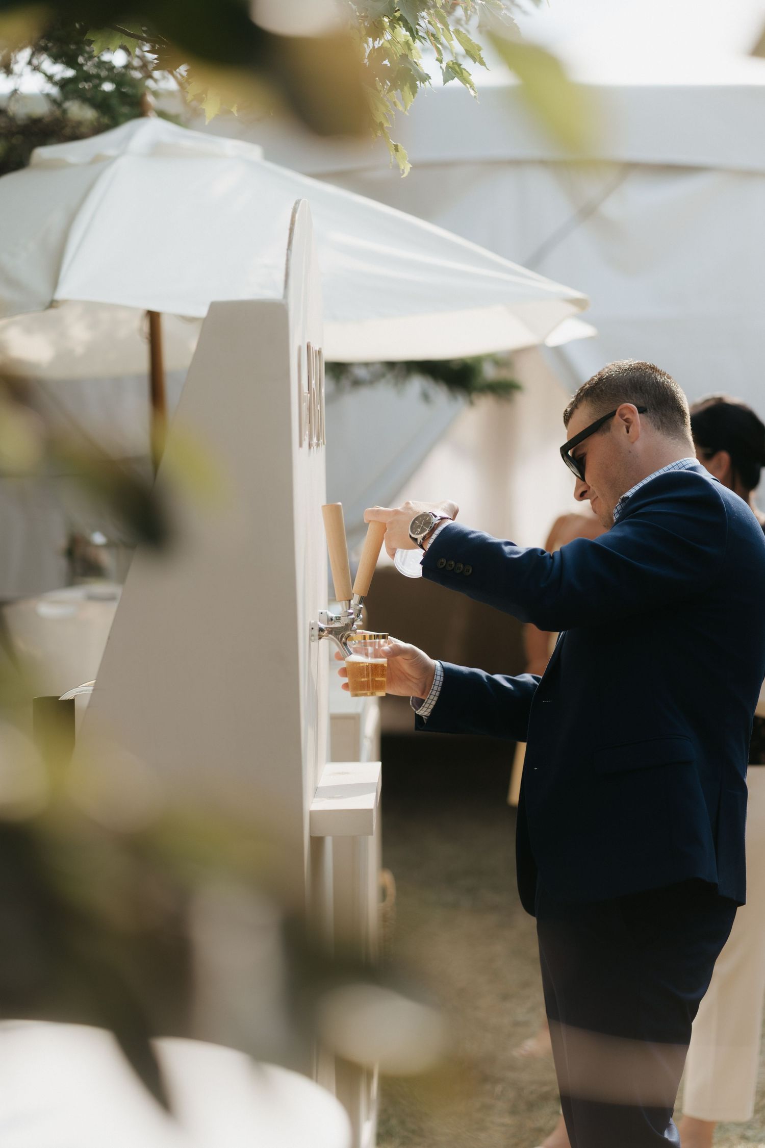 Someone in a navy jacket pouring drinks through a white wall partition at an outdoor event.