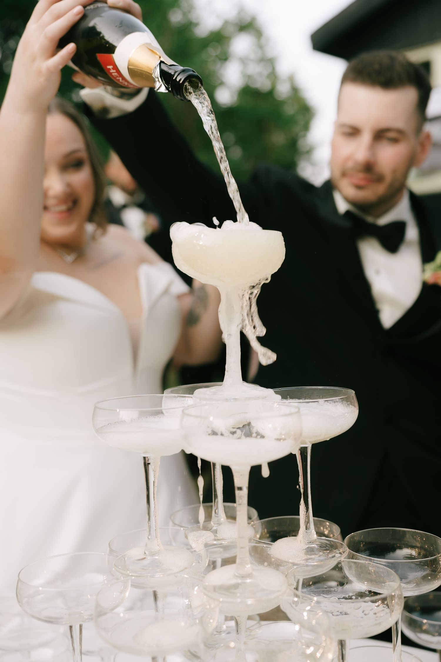 Clear champagne cascades down a tower of delicate coupe glasses in an outdoor setting.
