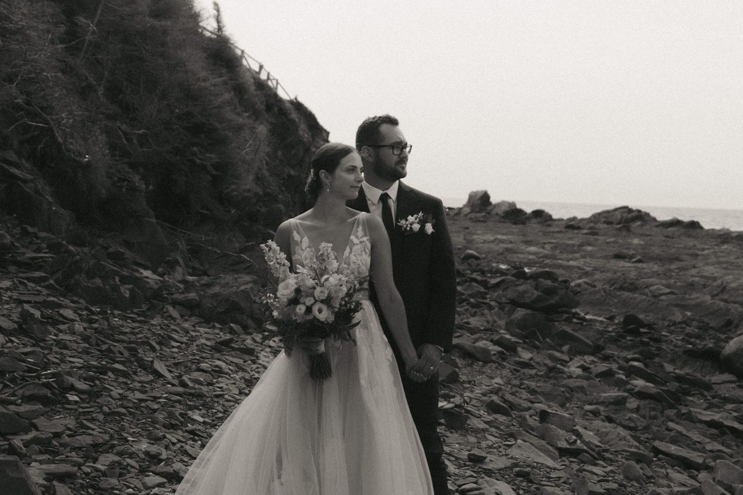 Elegant couple poses together for romantic black and white wedding photos on a rocky cliff by the ocean.