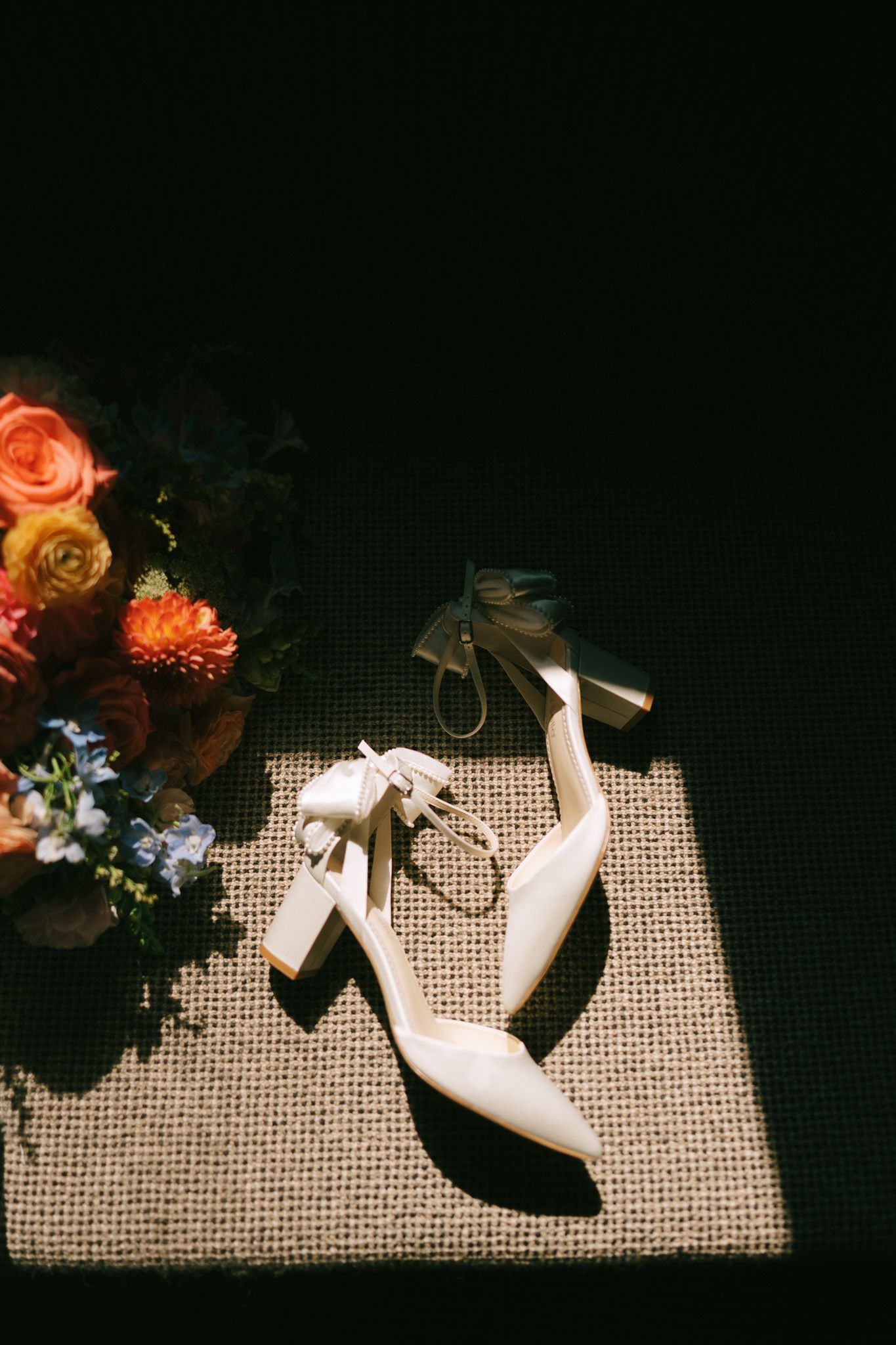 Classic white heels and bright floral bouquet arranged on woven material with dramatic shadows.