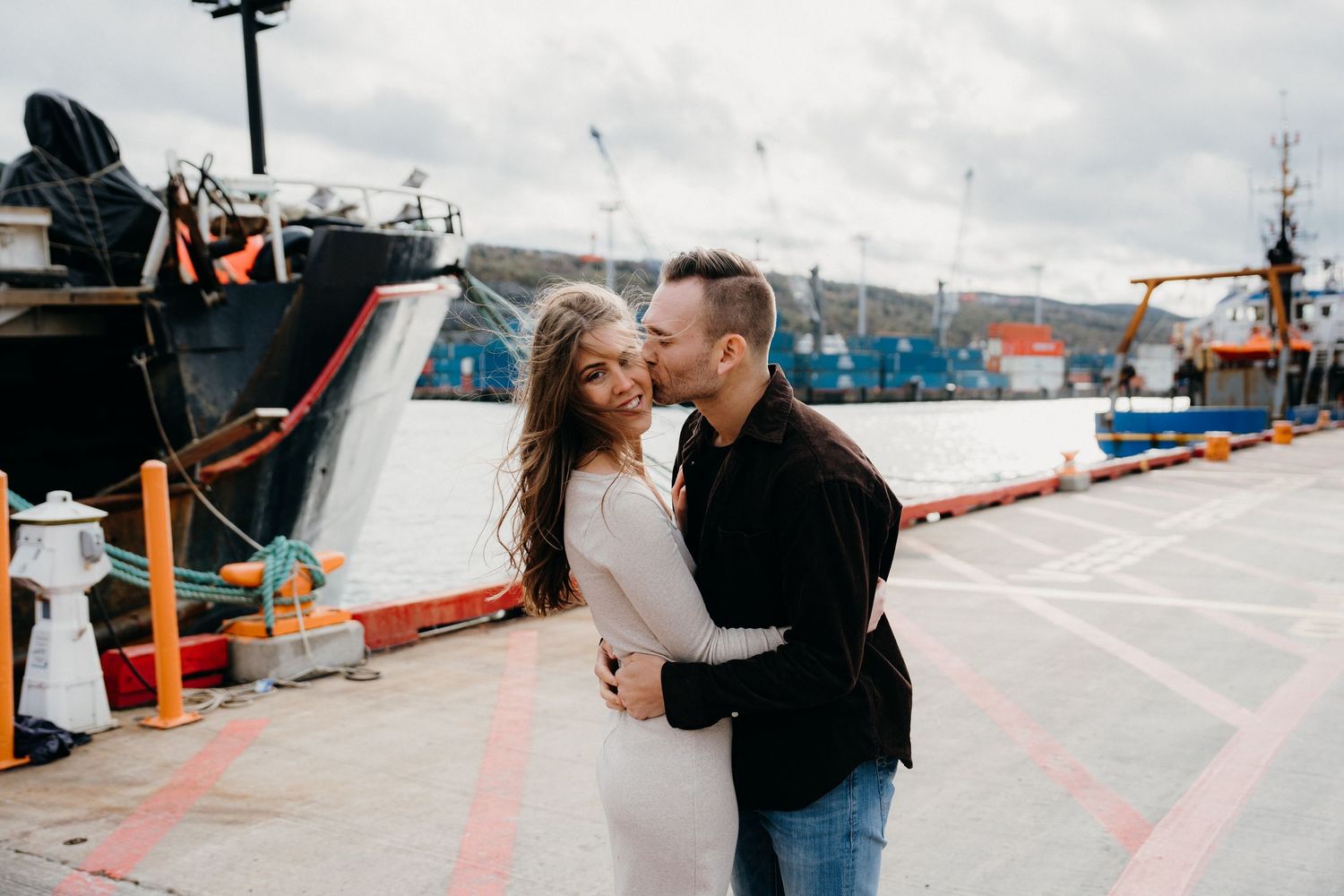 Two people share an intimate moment on a waterfront with boats and harbor equipment visible.