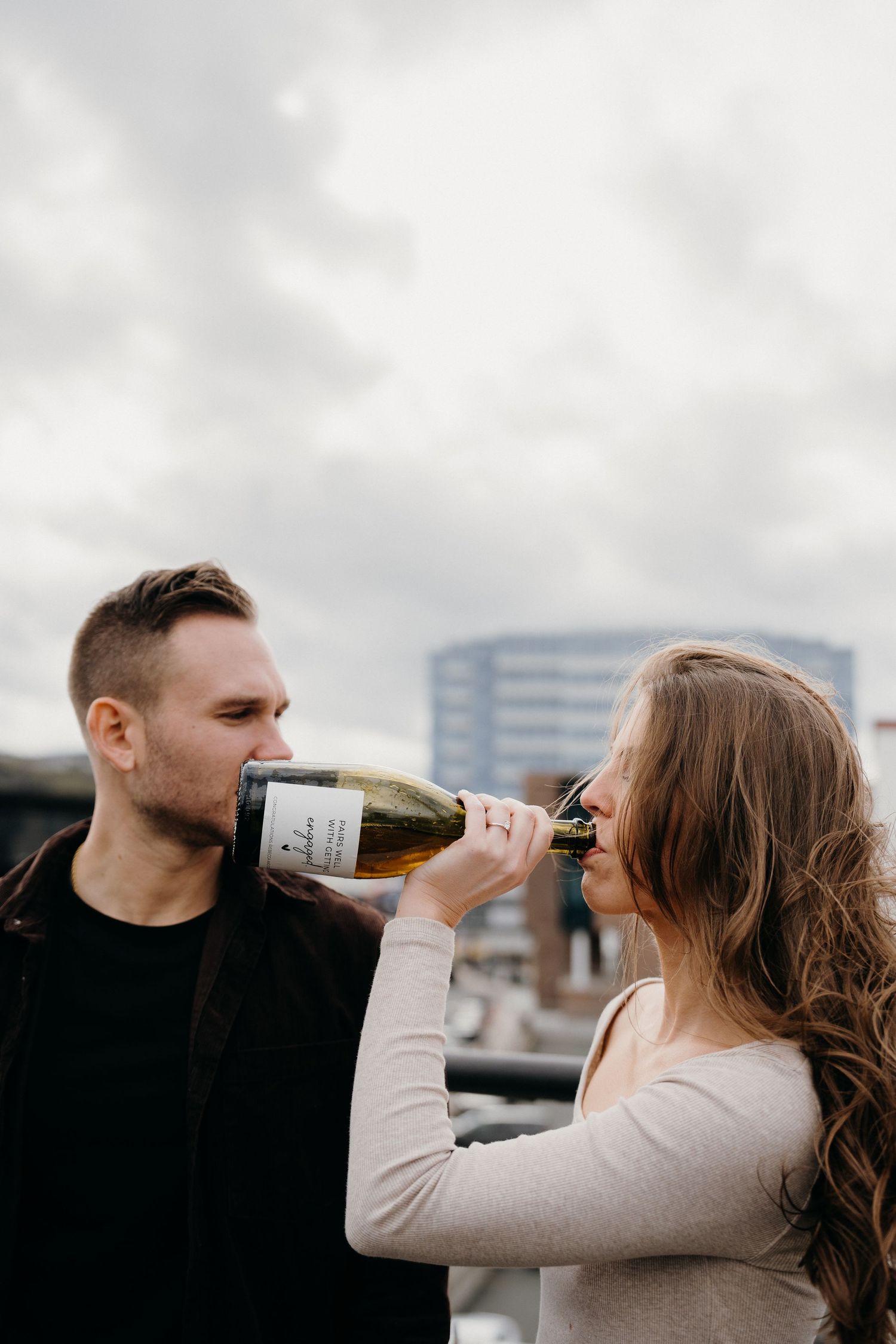 Two people drink from bottles while standing close together on an urban rooftop.