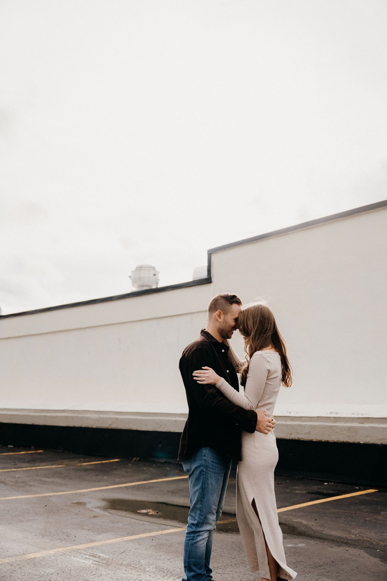 Two people embrace in a minimalist parking area with white walls creating a stark backdrop.