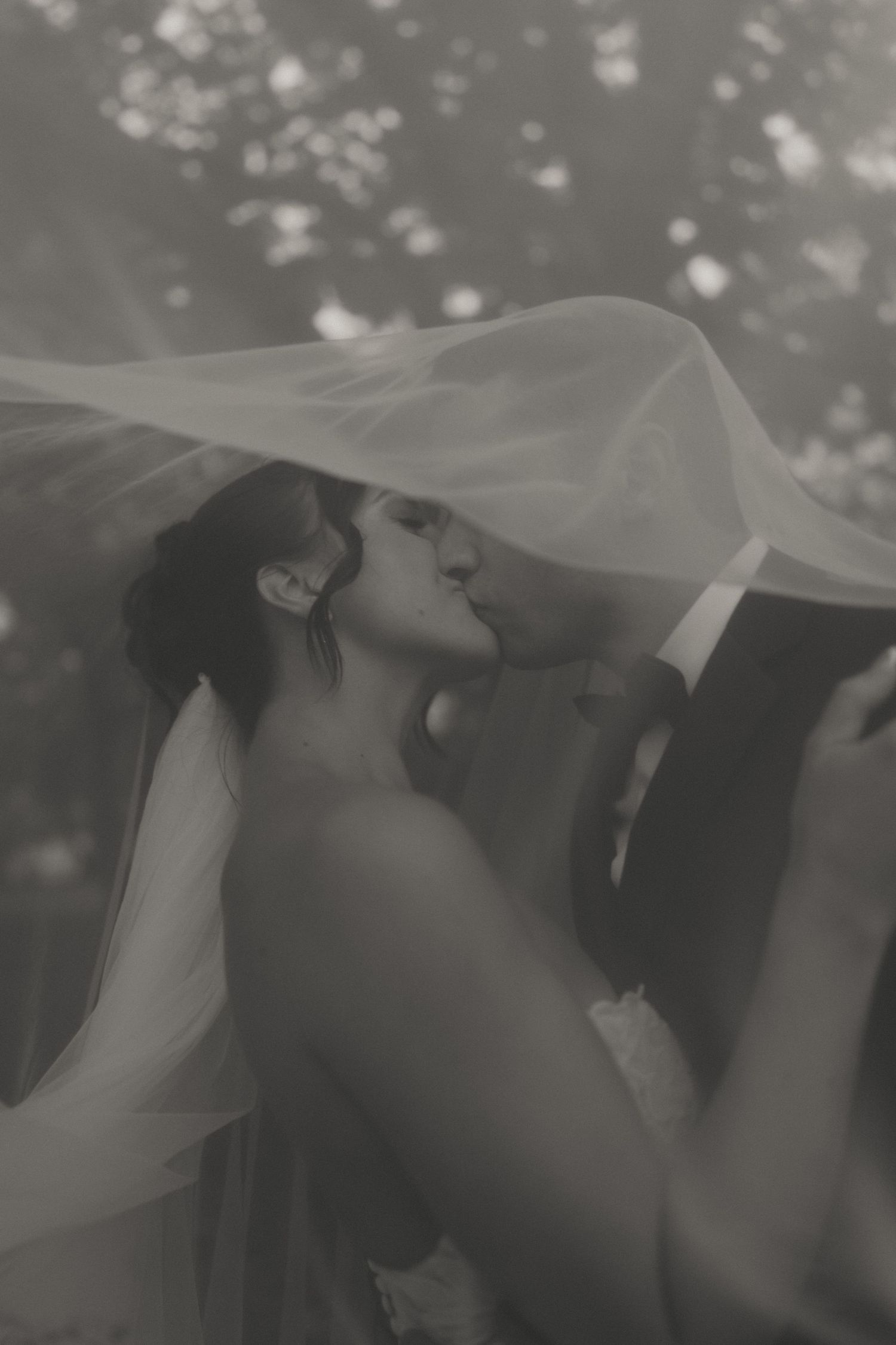 Black and white wedding photo of couple kissing under a flowing veil outdoors.