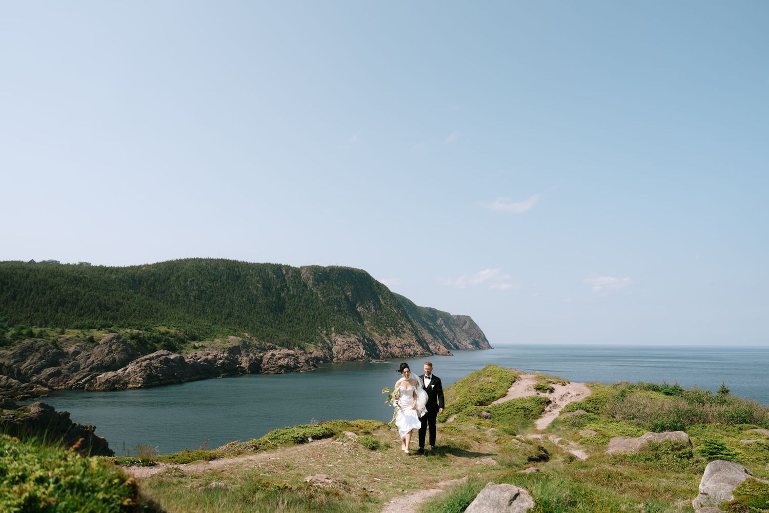 Couple in wedding attire standing on rocky coastal cliffside overlooking scenic ocean inlet with lush green hills.