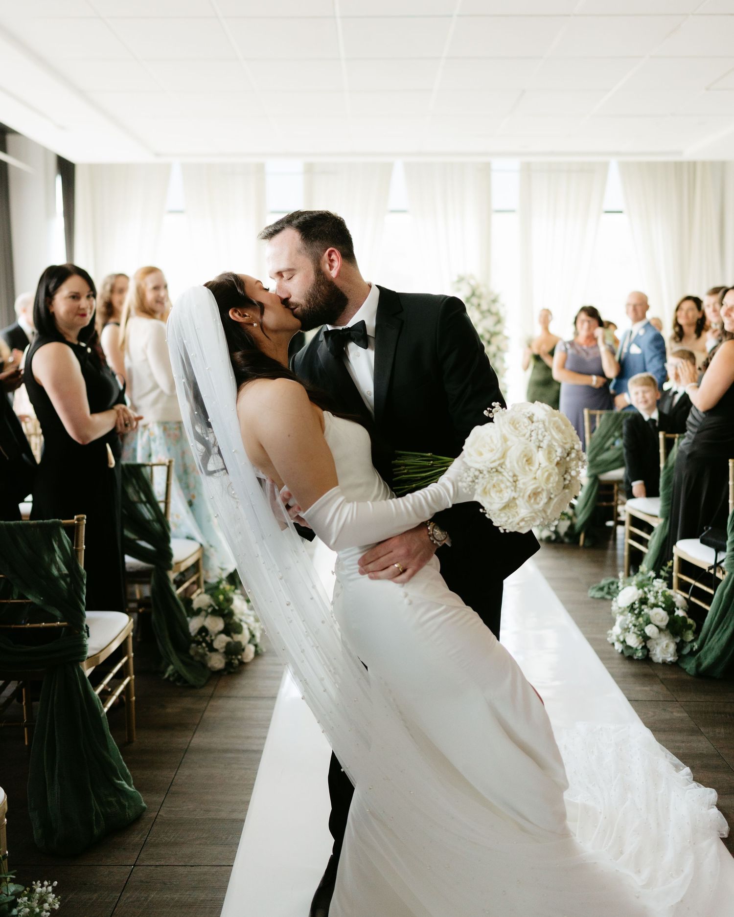 Bride and groom share a romantic dip kiss at wedding ceremony aisle, bride in white gown with veil holding bouquet.