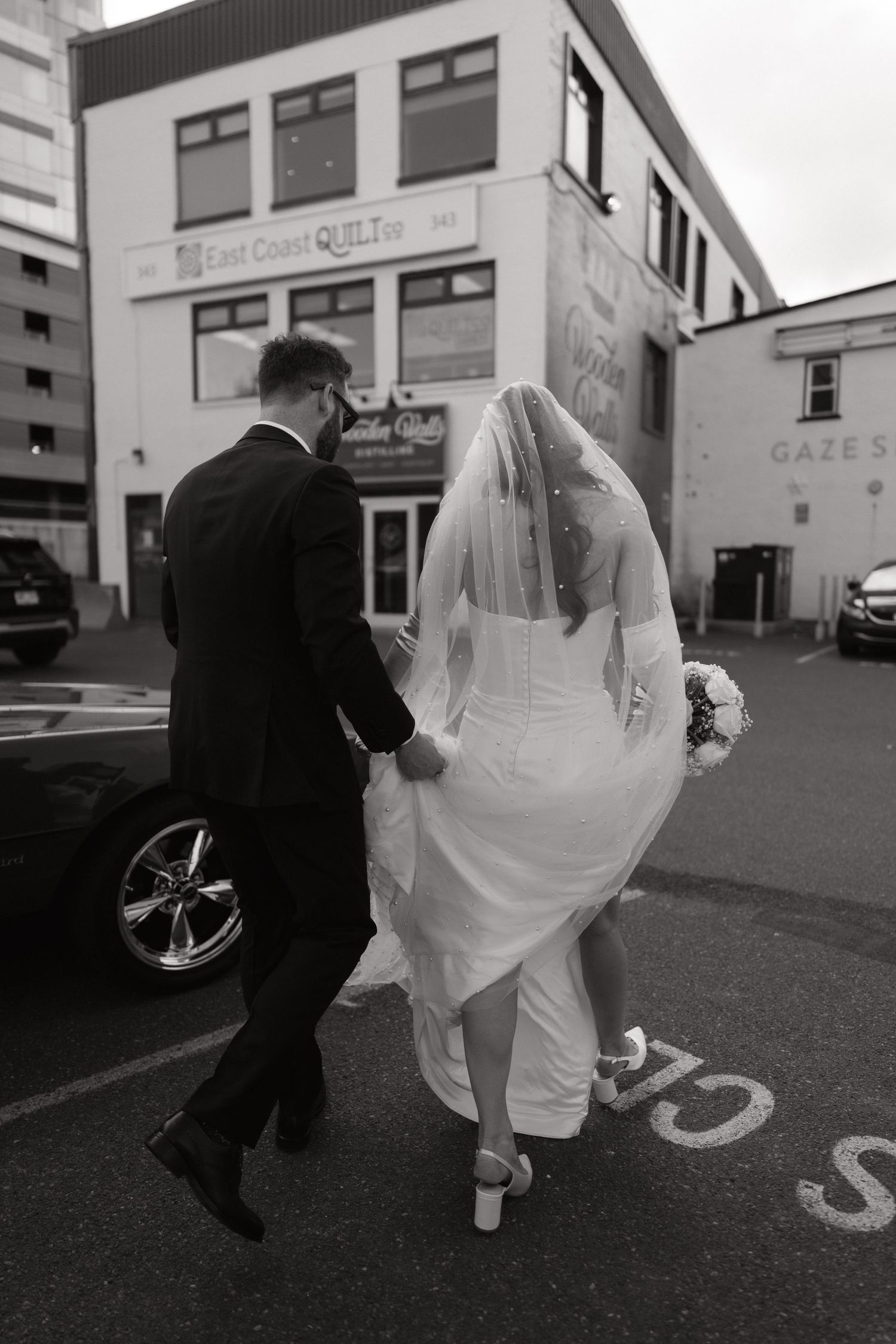 Black and white photo of a bride and groom walking through a parking lot, bride holding bouquet and veil flowing.