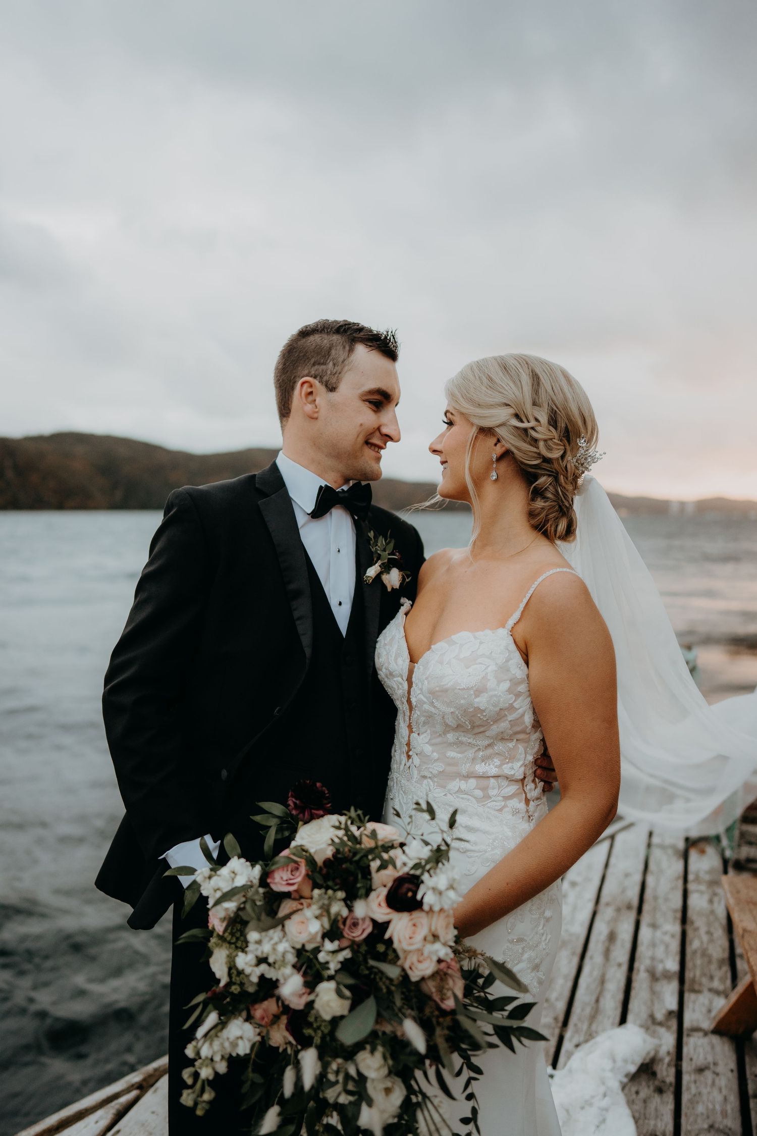 A romantic wedding couple embraces on a coastal pier at sunset with elegant floral bouquet details.
