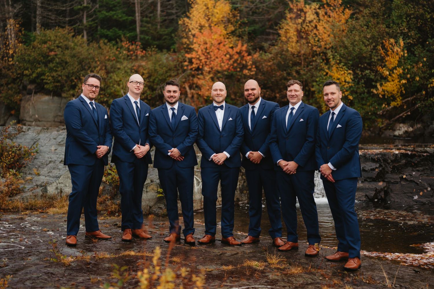 A group of seven men in navy suits stand together outdoors against autumn foliage and rocky terrain.