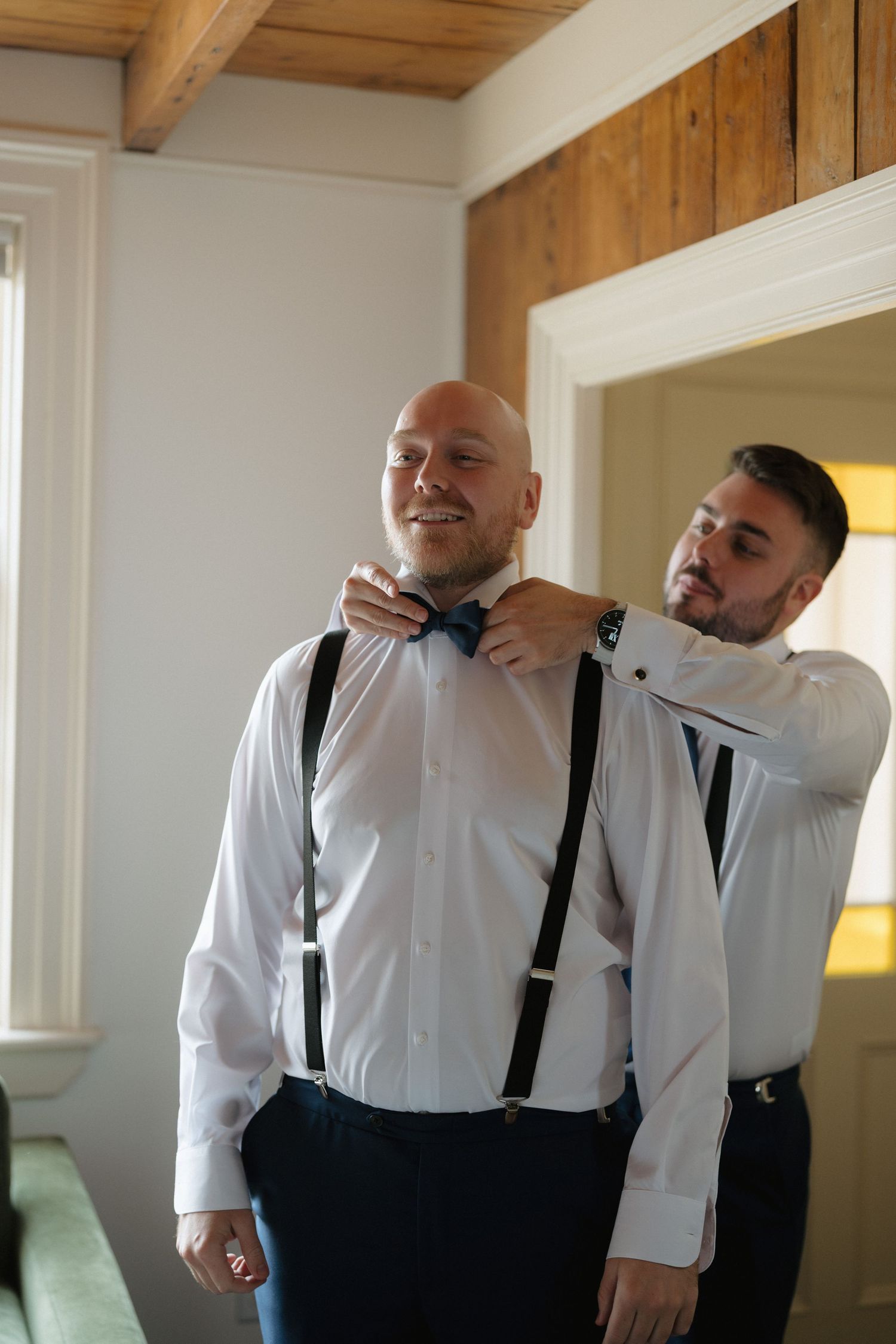 Groomsmen in white shirts and black suspenders help each other get ready before a wedding ceremony.