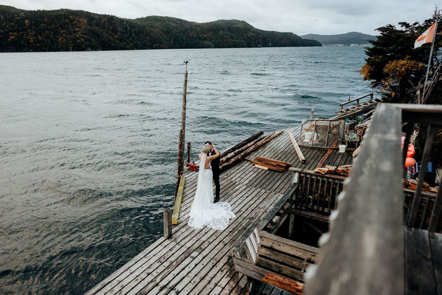 Wooden dock stretches over dark rippling waters with mountains and cloudy skies in the background.