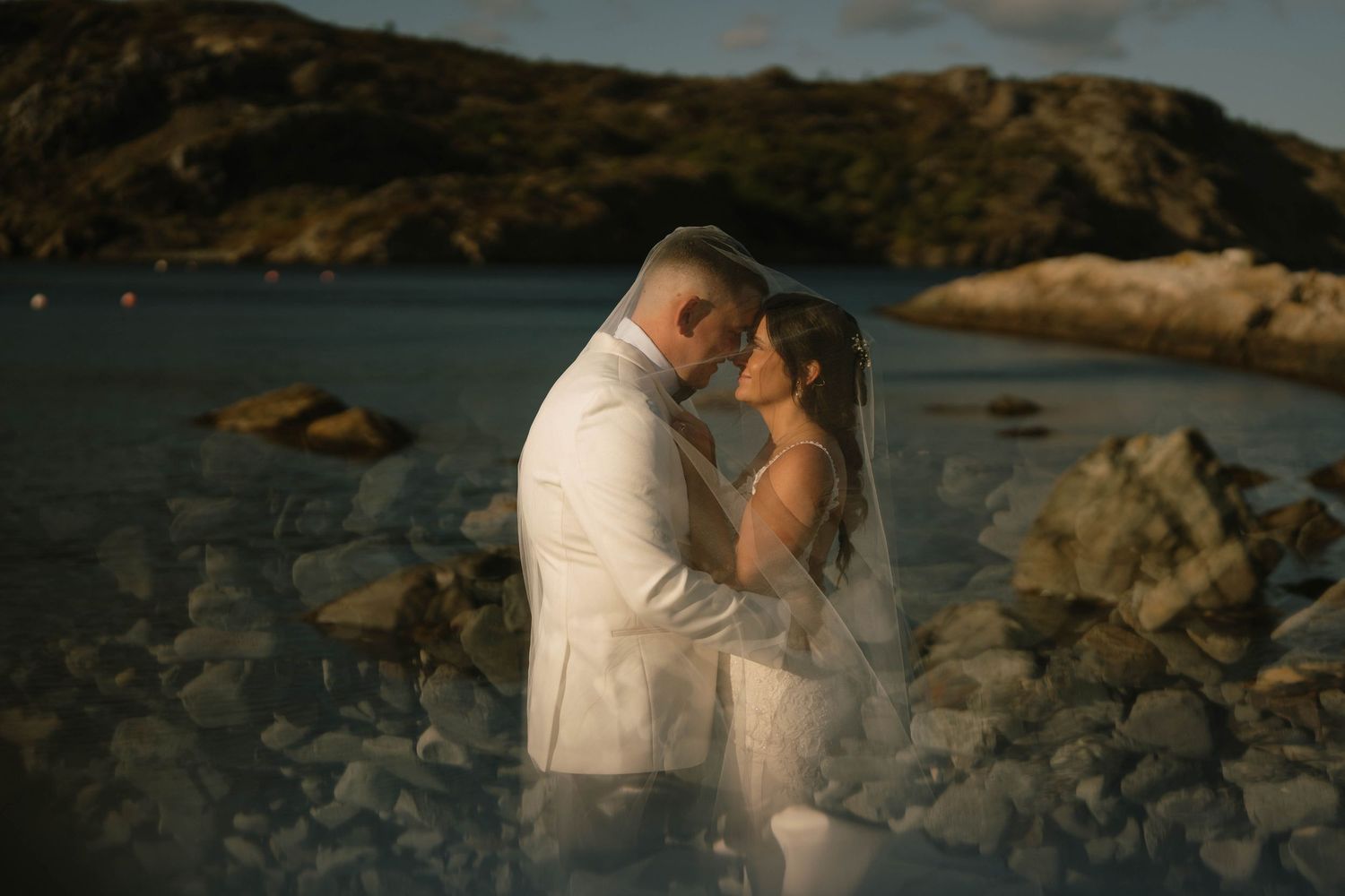 Romantic coastal photoshoot at sunset with couple embracing among rocky cliffs by the ocean.