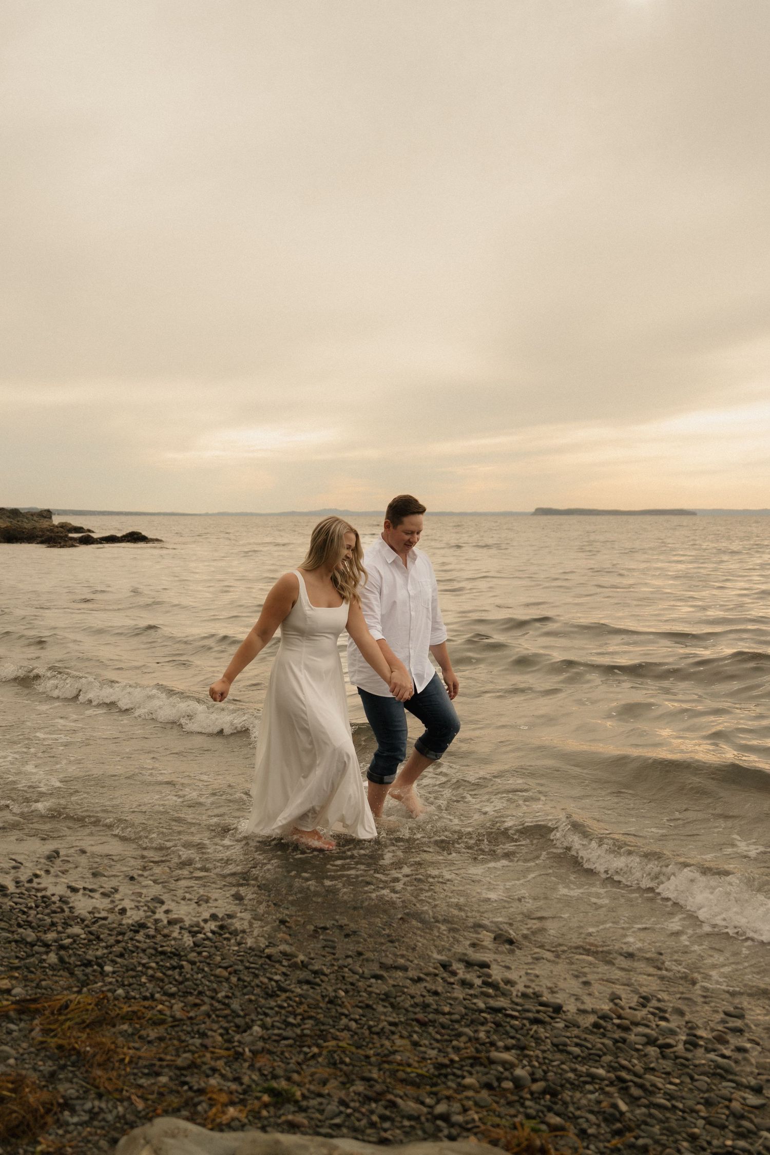 A couple in wedding attire playfully explores a rocky beach at sunset along the ocean shore.