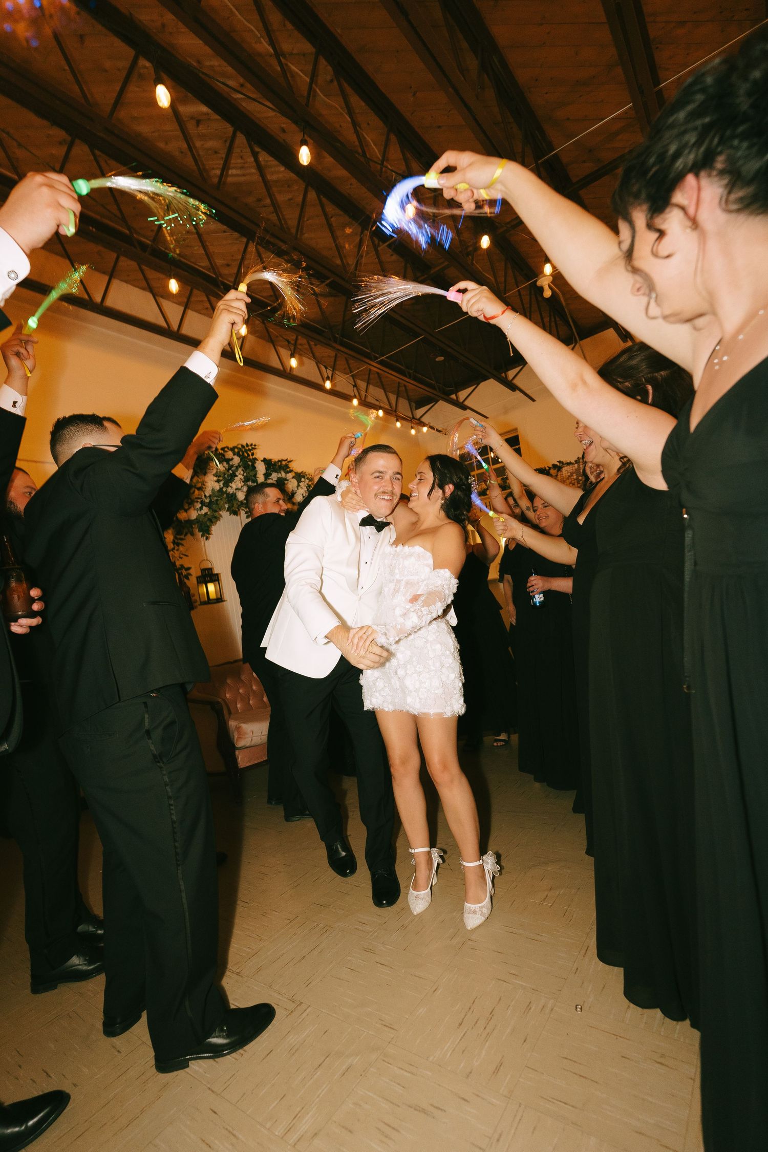 Wedding guests in black suits dance and celebrate on the dance floor as a person jumps energetically in white attire.