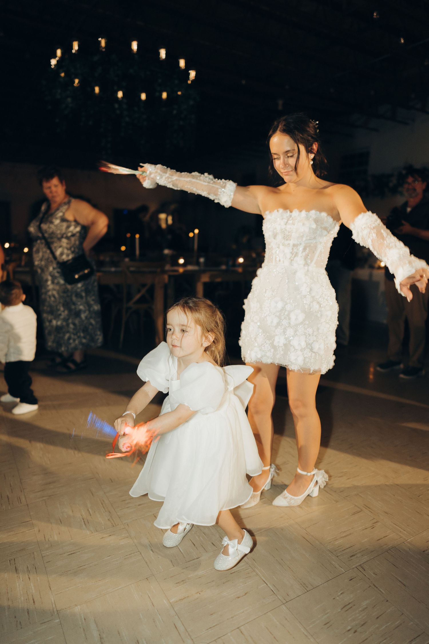 Dancers in white dresses perform energetic moves on a dance floor under dim lighting and string lights.