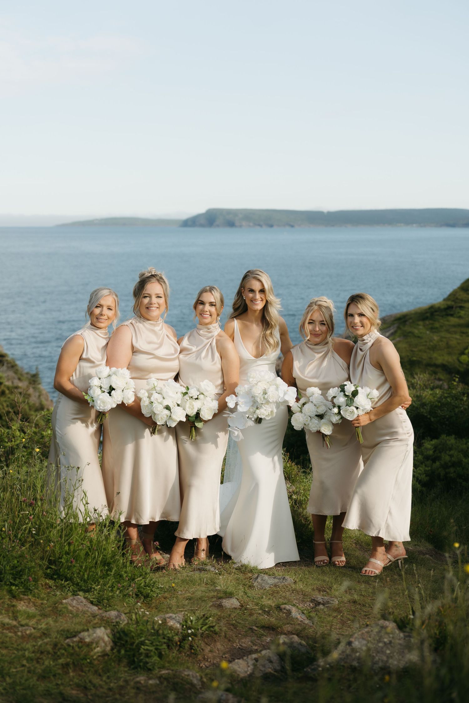 Bridal party in light-colored dresses poses against coastal backdrop holding white bouquets.