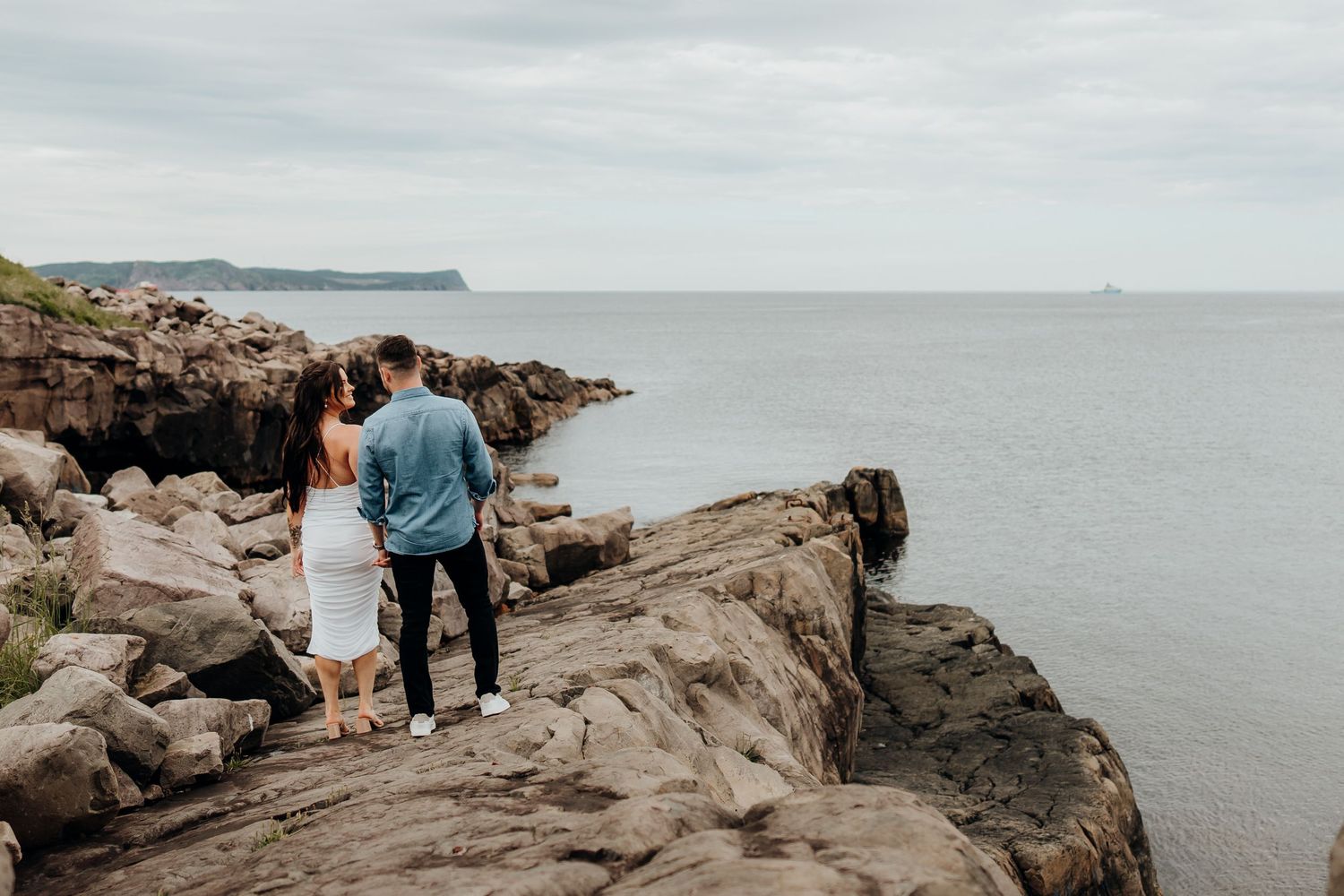 Two people walk hand in hand along rugged ocean cliffs with waves crashing below.