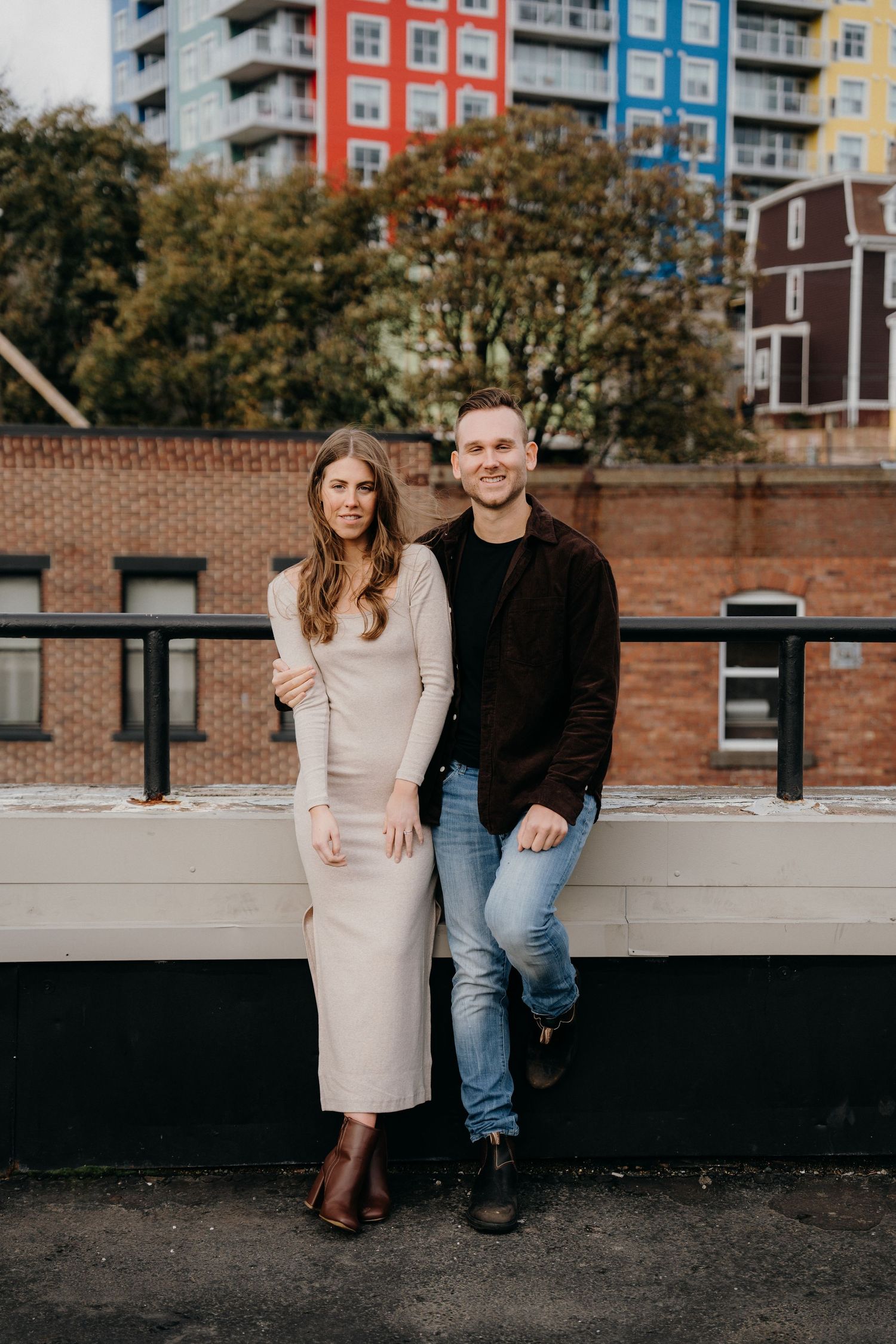 Couple in contrasting outfits posing together in front of an urban backdrop with colorful apartment buildings.