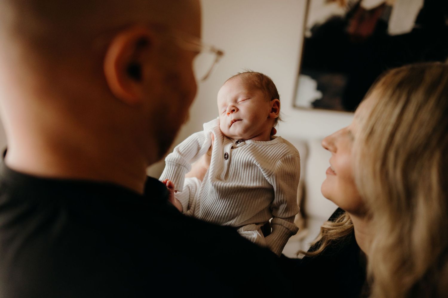 Parents share joyful expressions while holding their newborn baby in a bright living room.