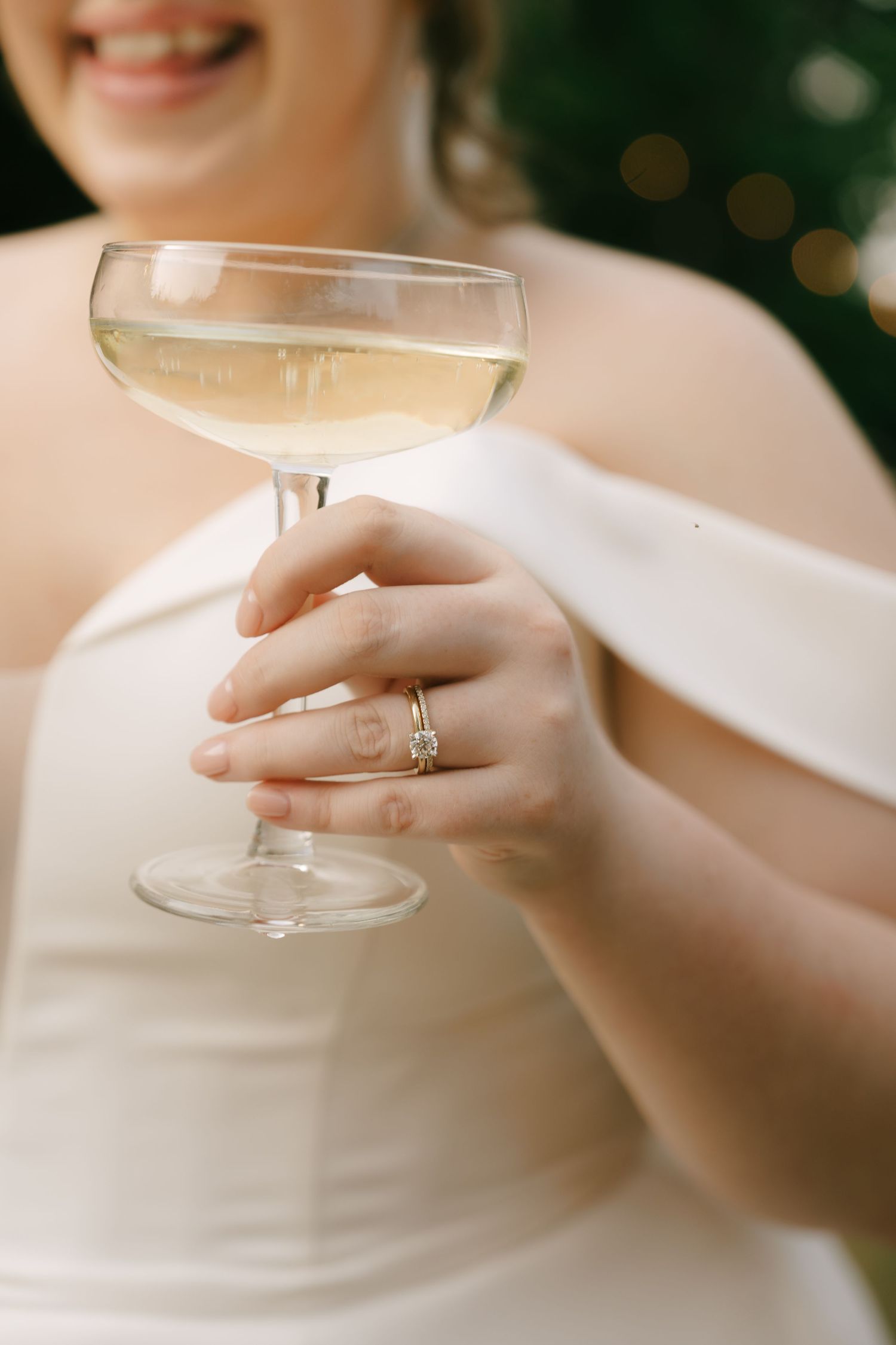 Close-up of a champagne glass held by hands wearing an engagement ring and white dress.
