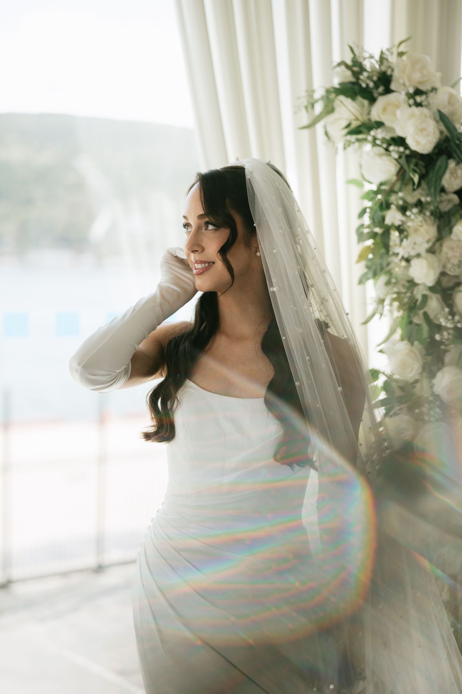 Elegant bride in white gown and veil stands by window with floral arrangement, rainbow light effect.