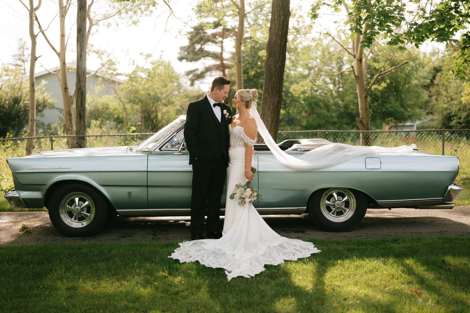 Bride and groom pose kissing beside a vintage blue convertible classic car outdoors on their wedding day.